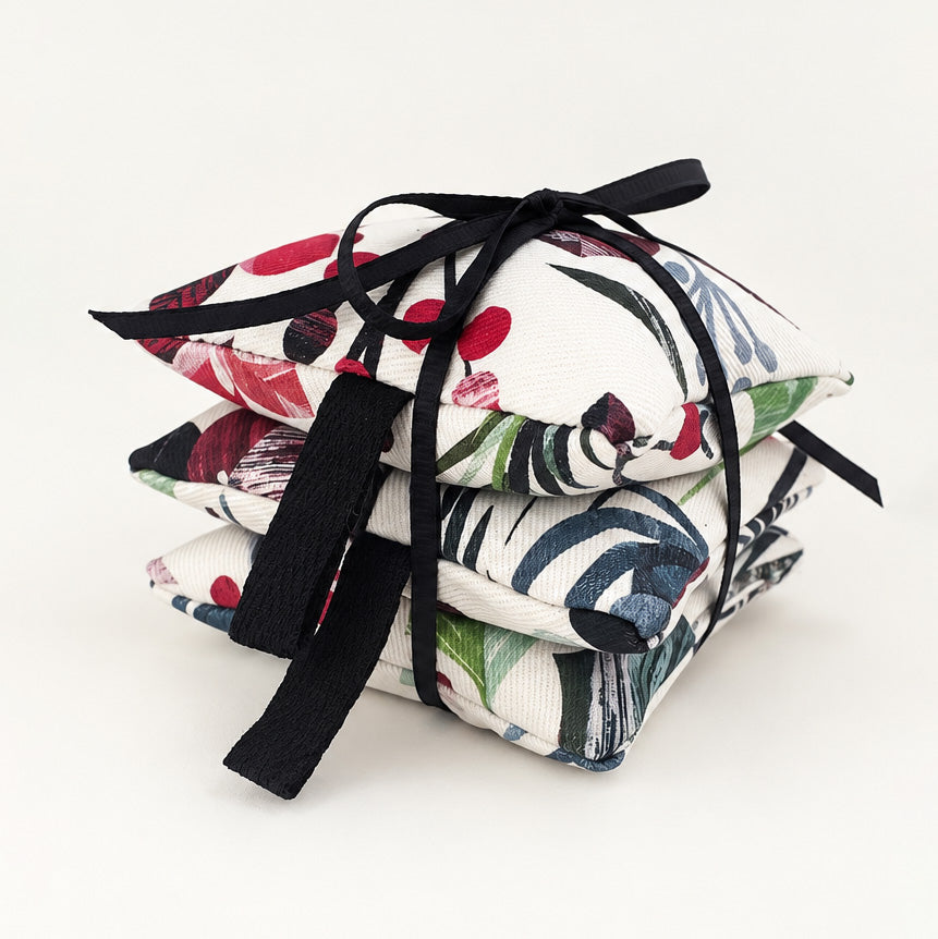 A pack of three square lavender bags with a white base in a colourful Christmas botanical print, tied together with a black ribbon bow on a clean white background.