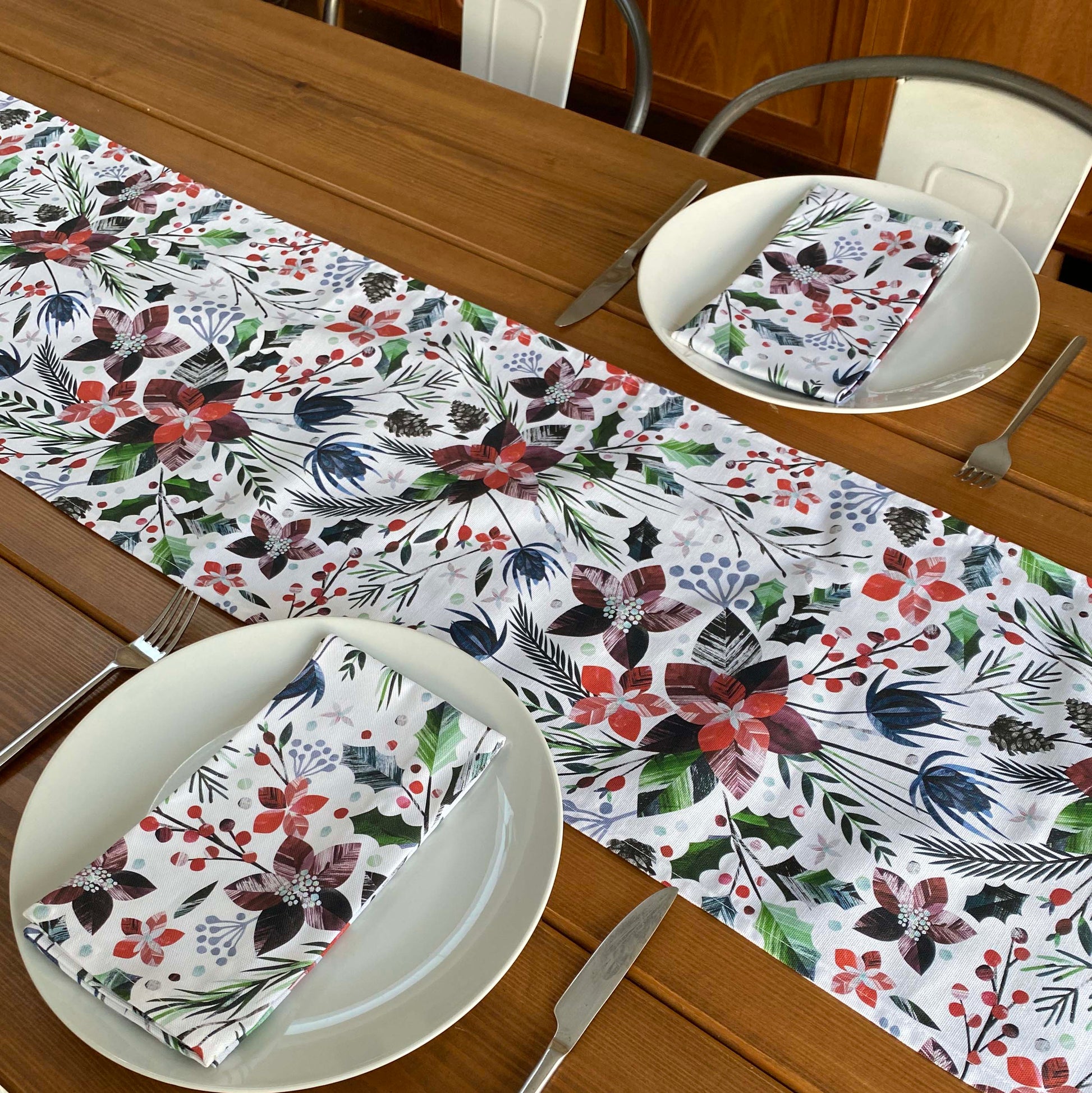 a Christmas Table Runner featuring red Poinsettia Flowers, green holly and red berries on a white background has been placed down the centre of a wooden table.  The table has been set for two with two matching napkins.