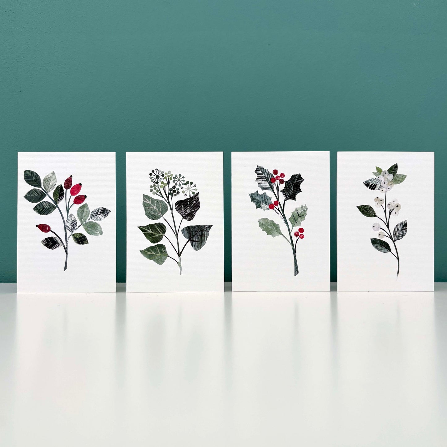 Four Winter Foliage arts cards featuring Rosehip, Ivy, Holly and Snowberry on a white background have been stood on a white shelf with a blue green background.