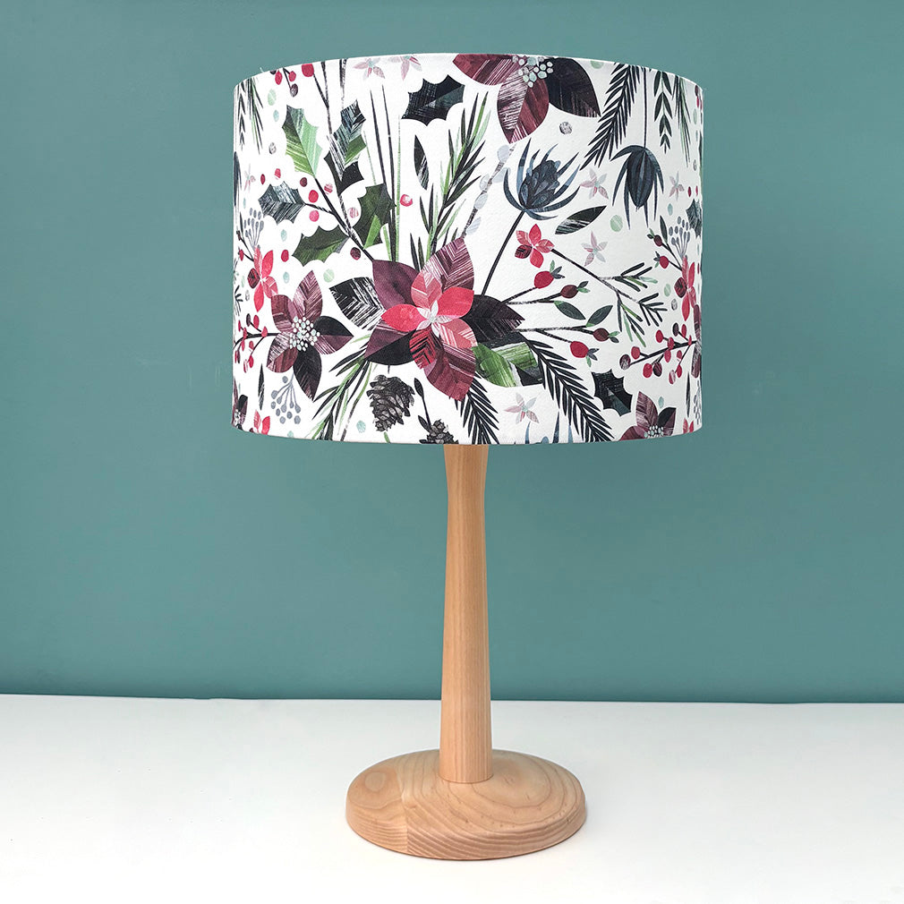 A medium Winter Floral Lampshade has been put on a wooden lamp on a white shelf with a blue green background.