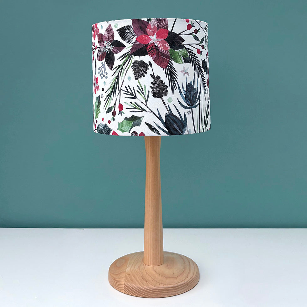 A small Christmas, winter floral patterned Lampshade has been put on a wooden Lamp base against a teal background.