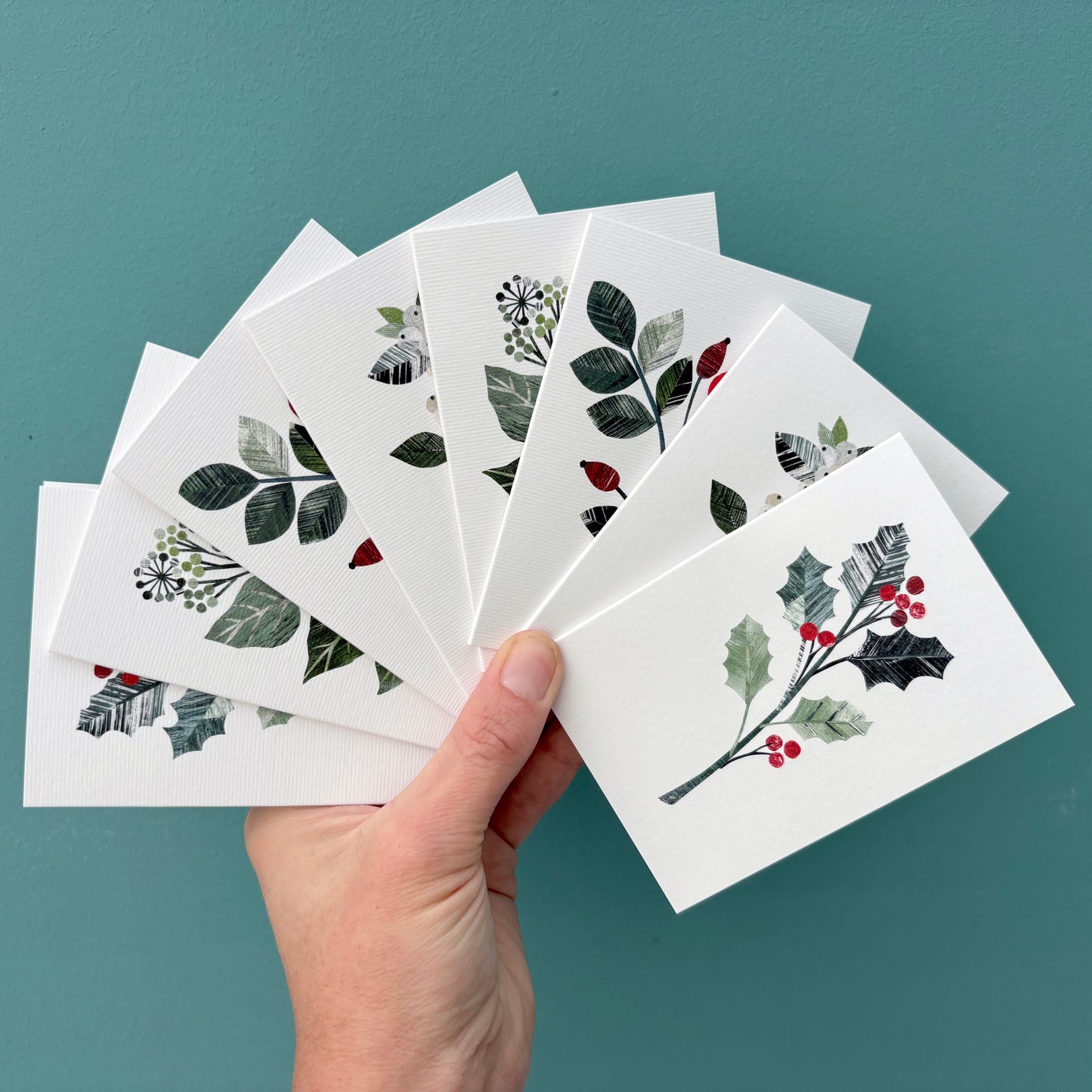 Hand holding eight mini cards with Winter floral designs on them against a teal background.