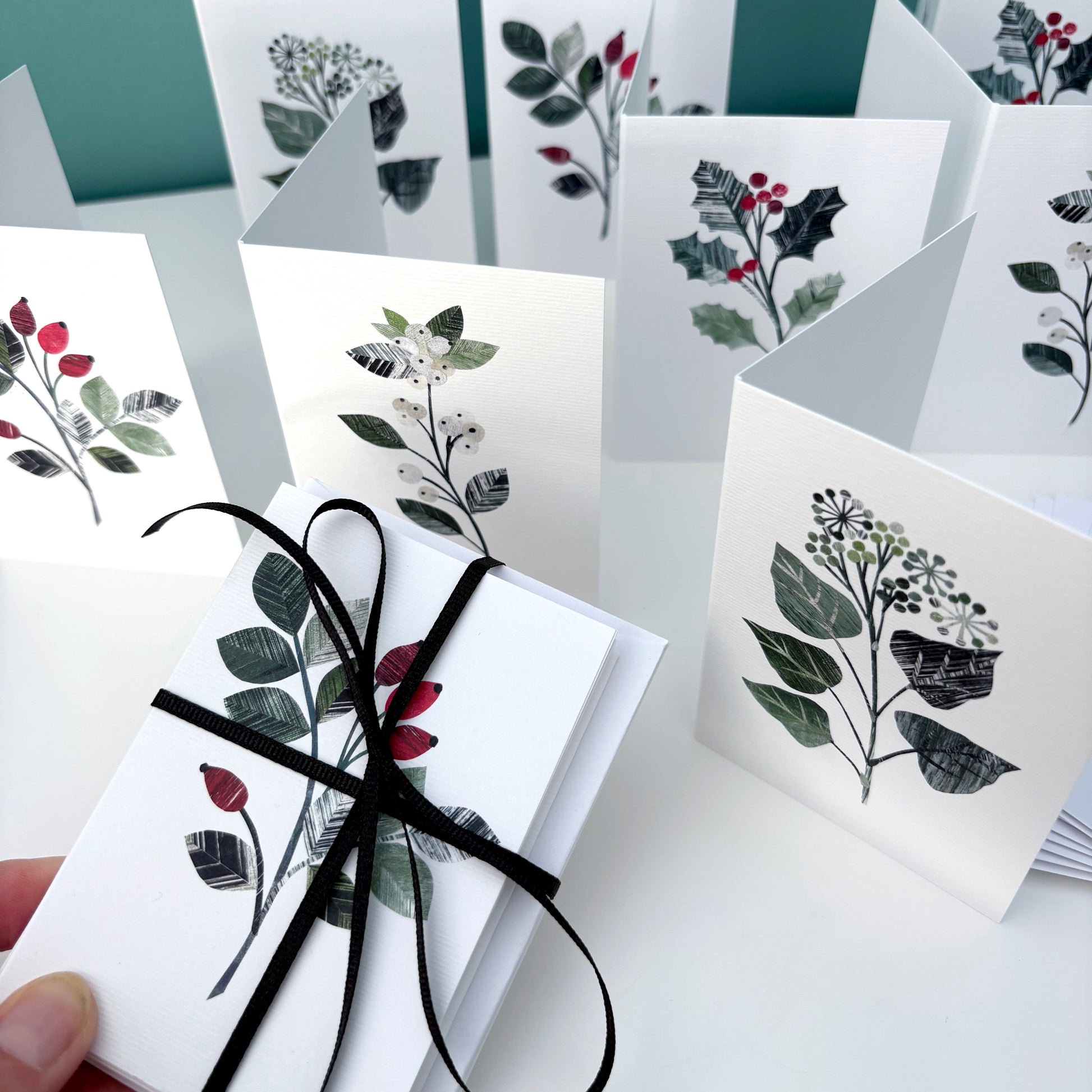 Set of Winter Floral greeting cards, 2 with a holly motif on them, two with Ivy, 2 with Rose hips and 2 with Snowberries on them.  At the front a pack of mini cards have been tied with black ribbon.