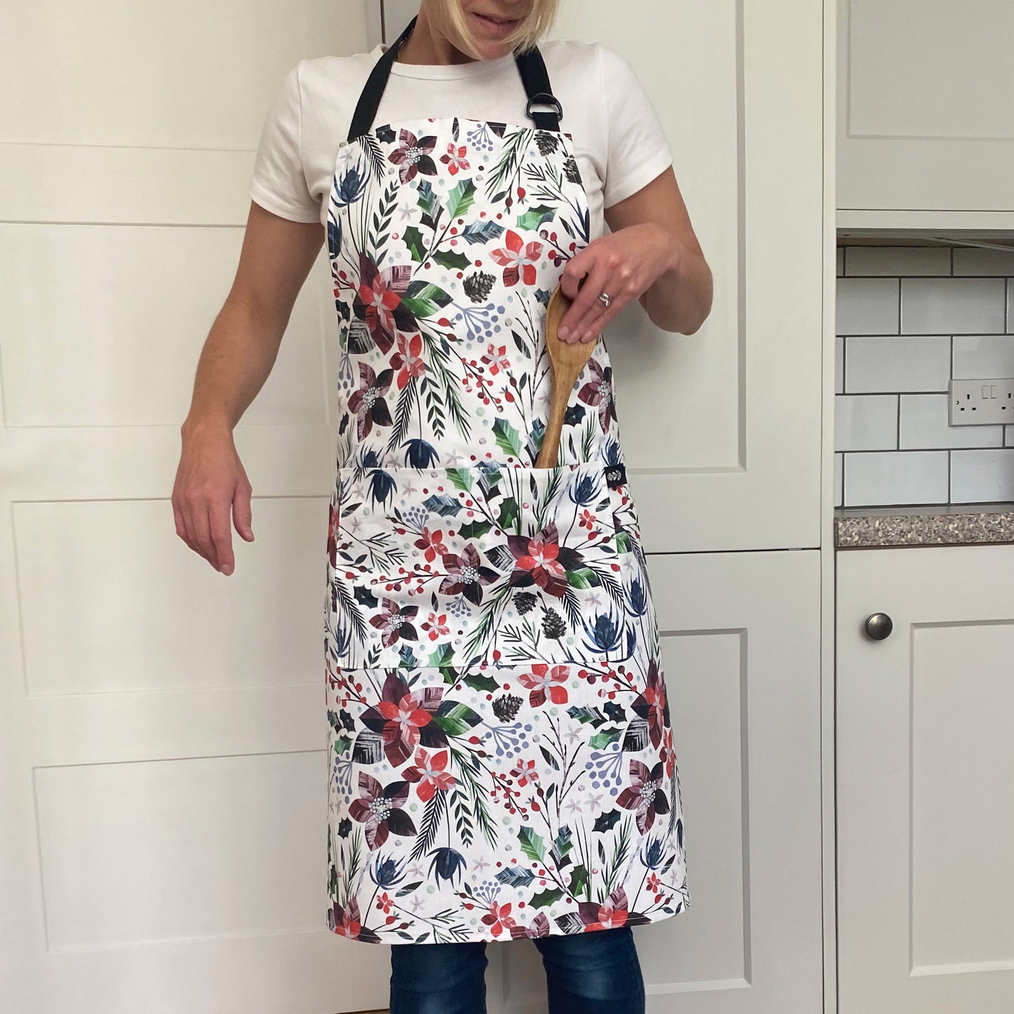 A lady is wearing the Winter Floral Apron with Red Poinsettia Flowers.  the Apron has two front pockets with a black adjustable neck strap.