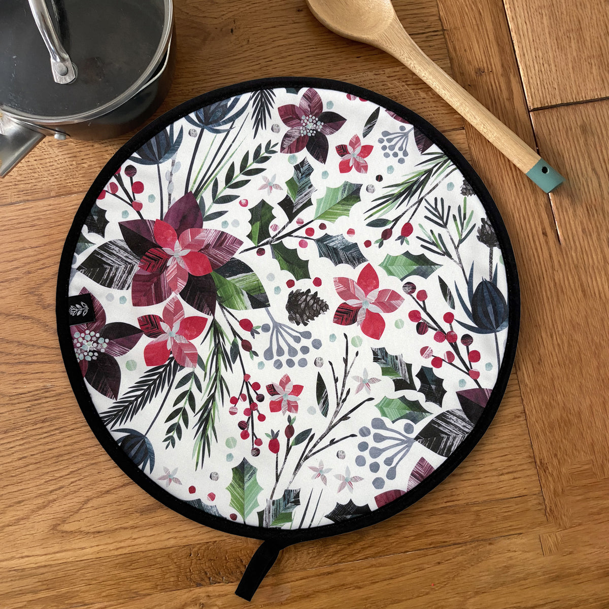 A round Aga Cover with a black trim and hanging loop.  Features a colourful winter botanical pattern of poinsettias, holly leaves, pinecones, and berries, shown on a wooden tabletop with kitchen props.