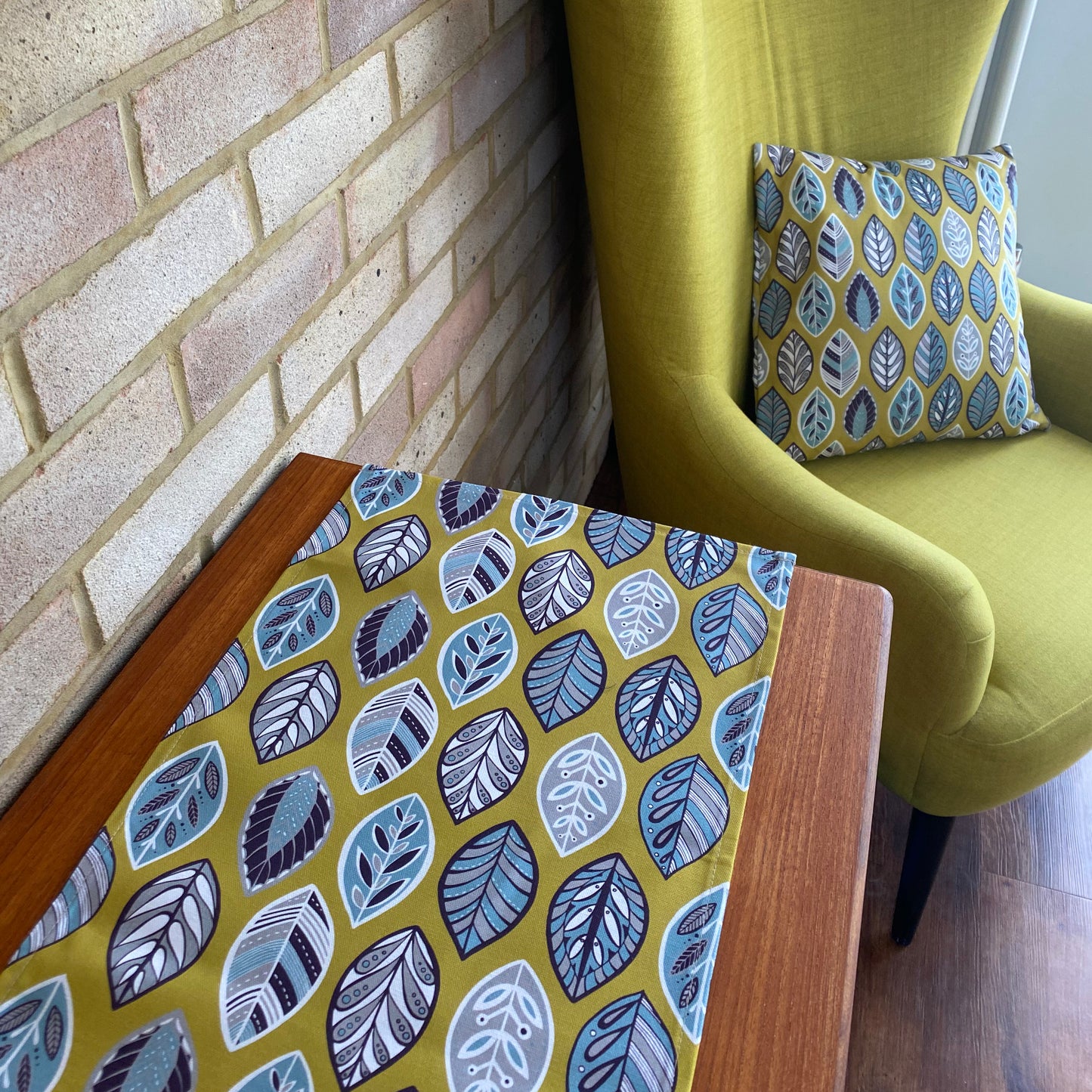 A yellow beech leaf table runner is shown here placed on a wooden side board.  There is also a matching cushion on a chair in the background.