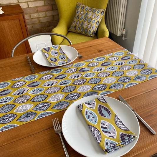 A yellow beech leaf table runner has been placed on the middle of a wooden table.  The table has been set for two and two white plates have matching napkins on them.