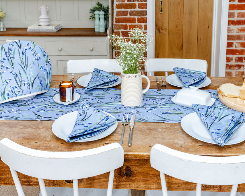 Dining table set with bluebell patterned table runner napkins, tea cosy, white plates, and a candle on a wooden floor.