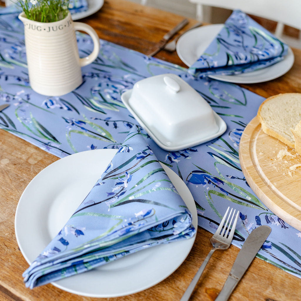 Dining table setting with bluebell floral runner and matching napkins, plates, and cutlery.