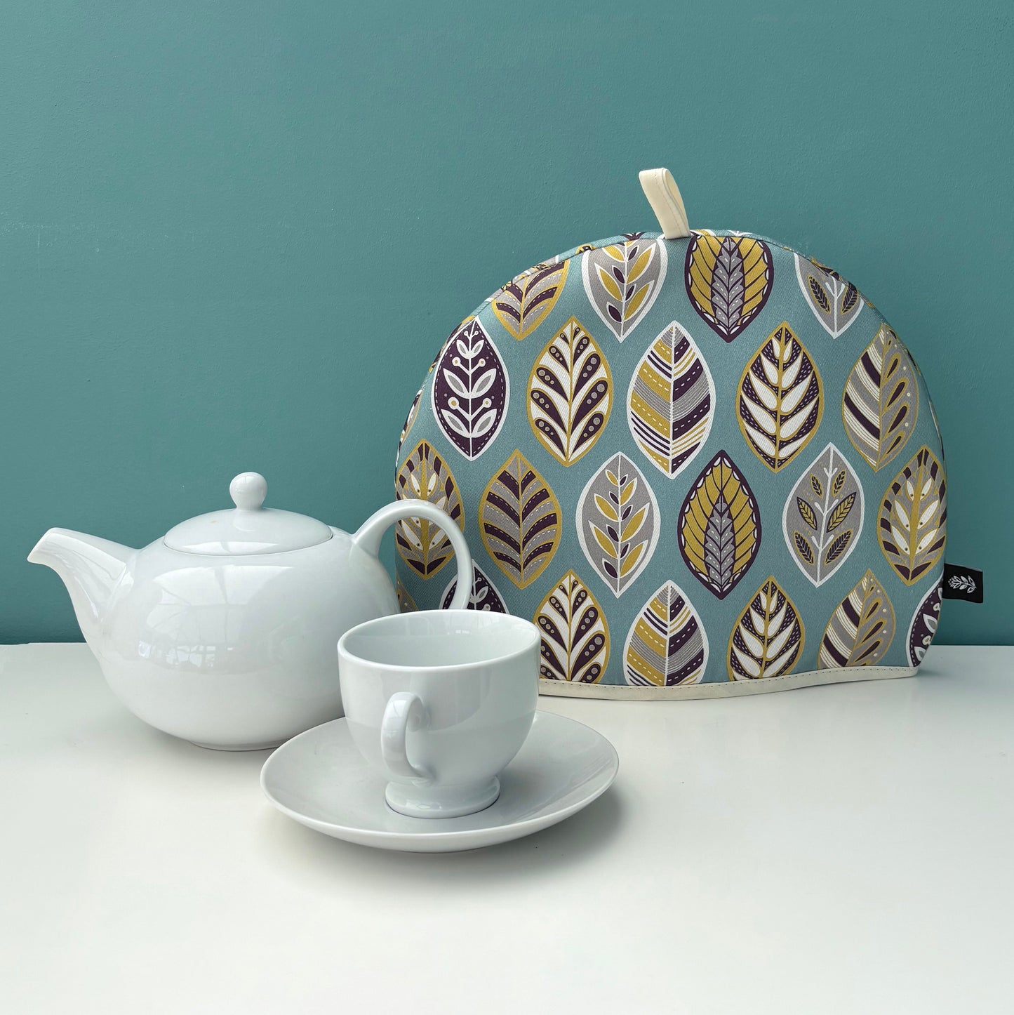 A Blue leaf tea cosy has been placed on a white shelf with a white tea pot and cup and saucer.