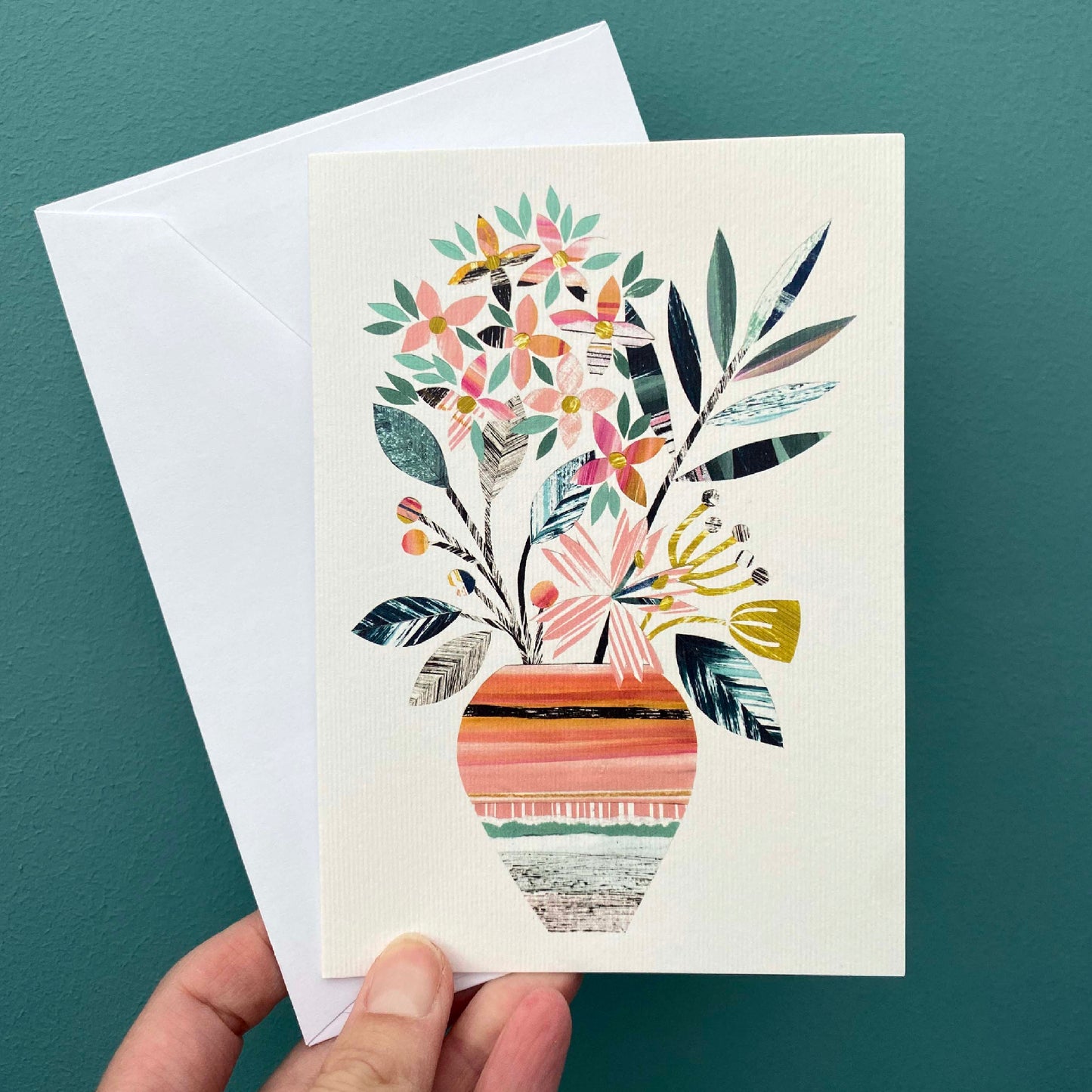 A white greeting card with a print of a colourful textured vase of flowers in green, orange, yellow and pinks, is being held with a white envelope in front of a blue green wall.