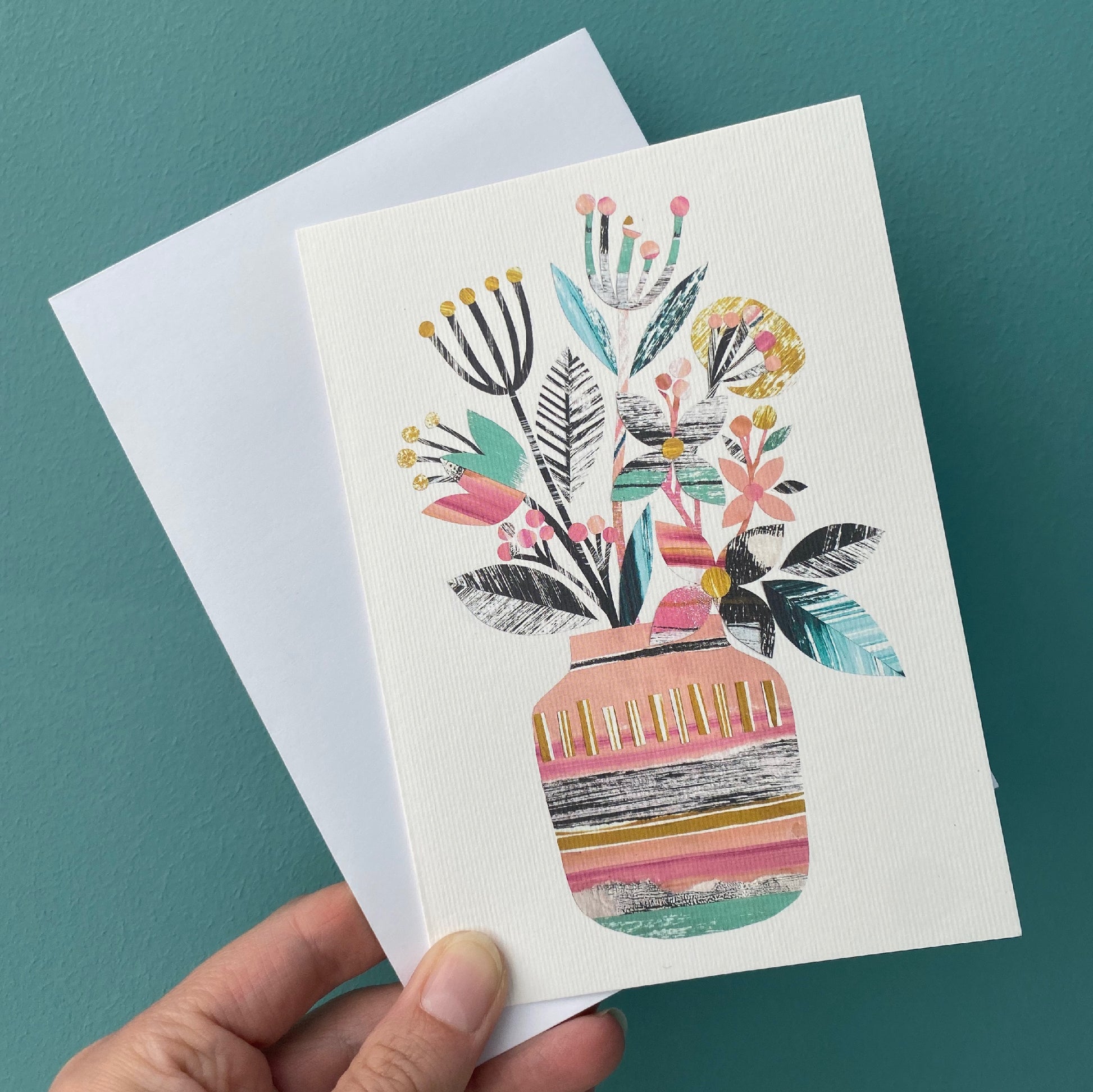 A colourful card with a vase of flowers on it is being held with a white envelope against a blue green background.