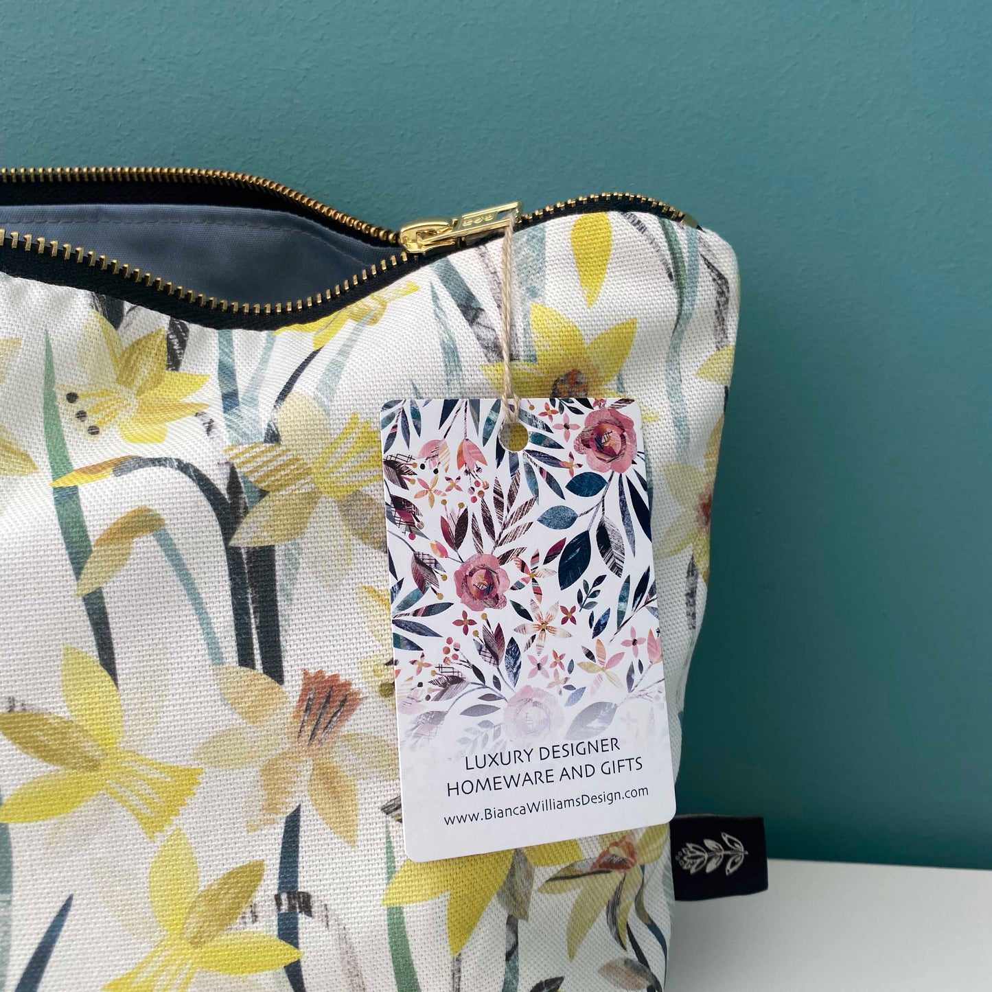 Close up of the Daffodil wash bag featuring yellow and orange Daffodil flowers with textured green leaves and stems on a white background.  A matching green lining can be seen on the inside of the bag.  The bags has a gold metal zip and a Brand label sewn into the outside seam.