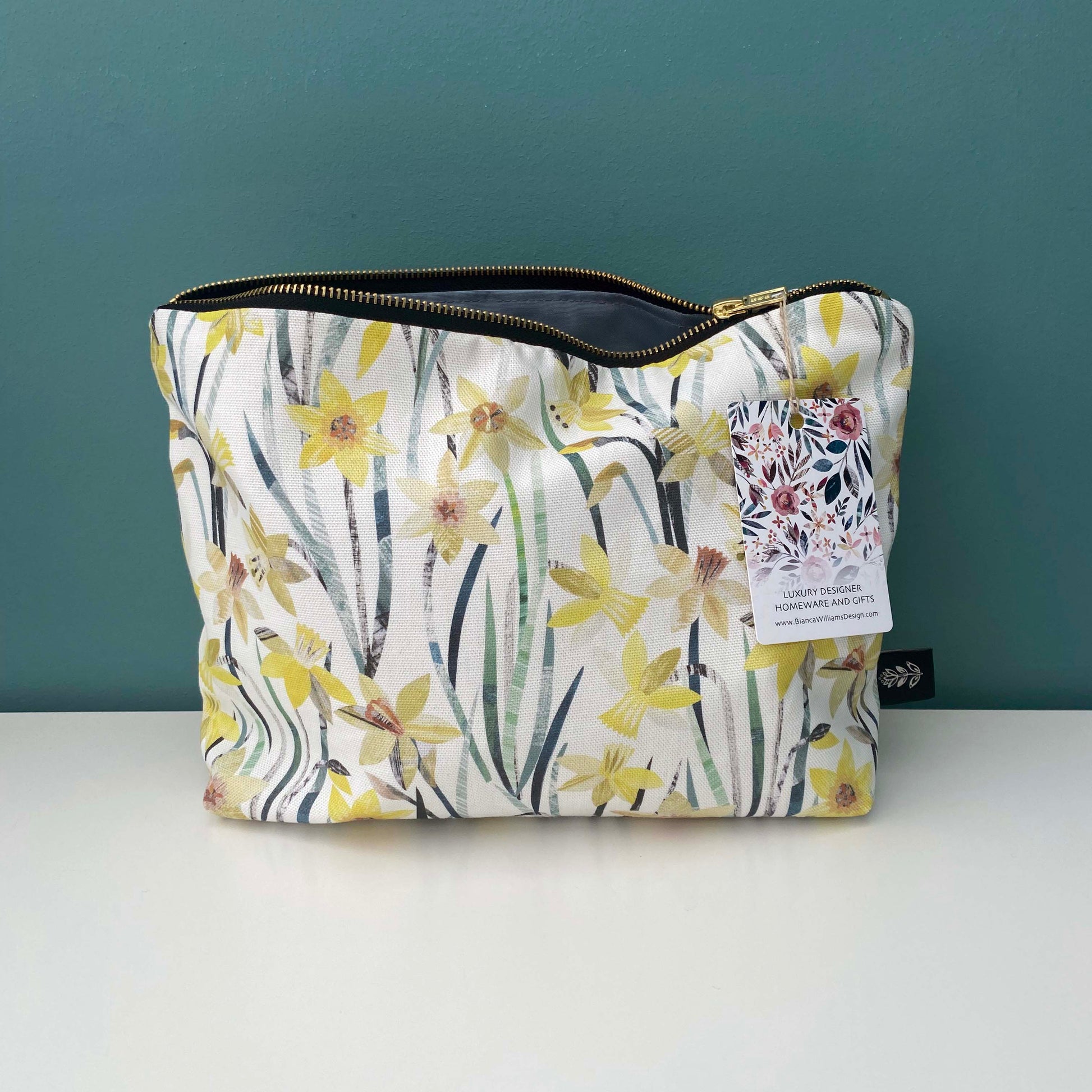 Large Daffodil Wash Bag, with Brand label sewn into the outside seam has been placed on a white shelf with a blue/green background.  The bag has a gold metal zip and a matching Green lining.