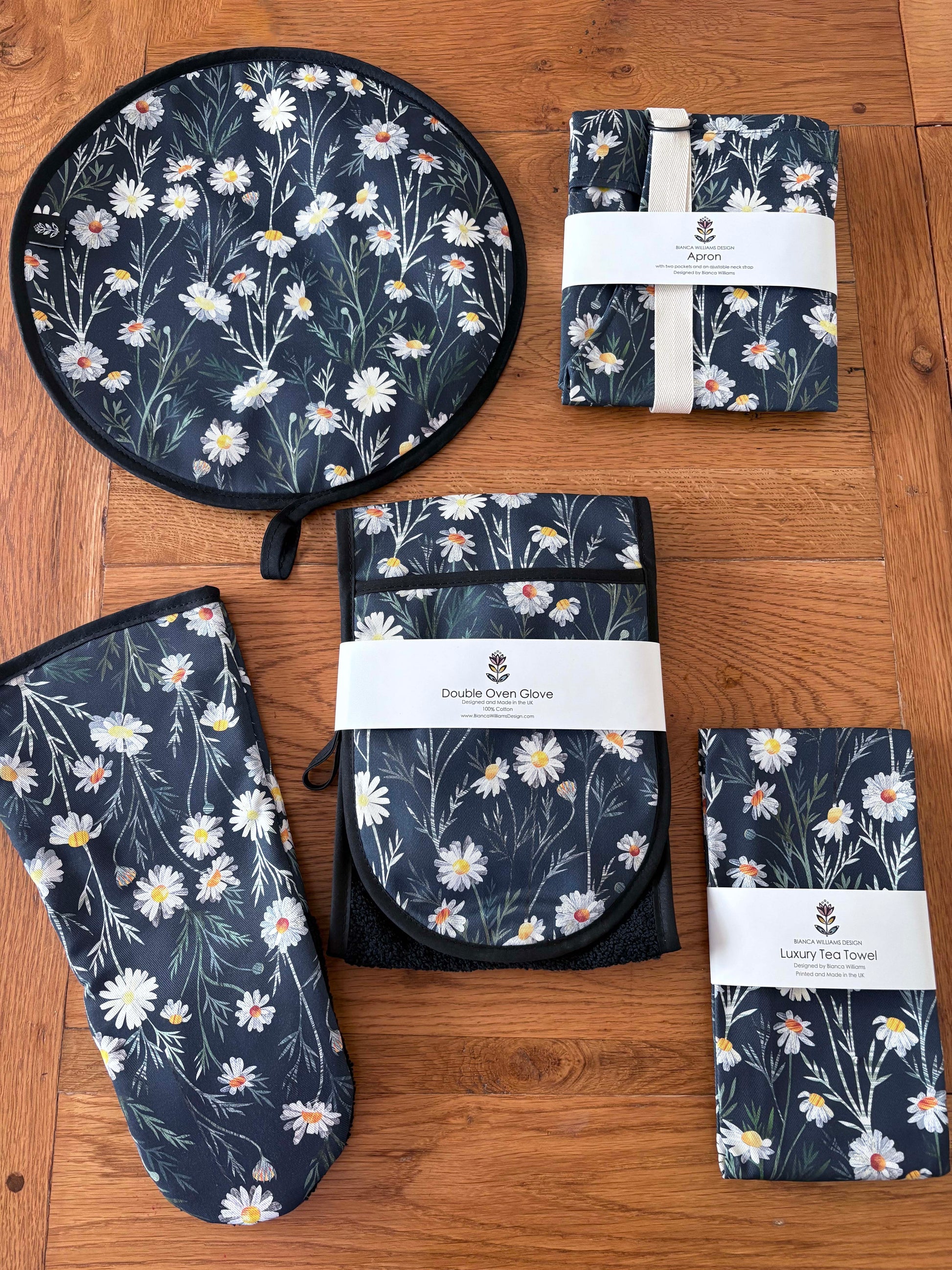 Set of Navy Daisy floral kitchen items including Double Oven Gloves, Gauntlet, Tea towel, Apron and an Aga Cover have been placed on a wooden surface.