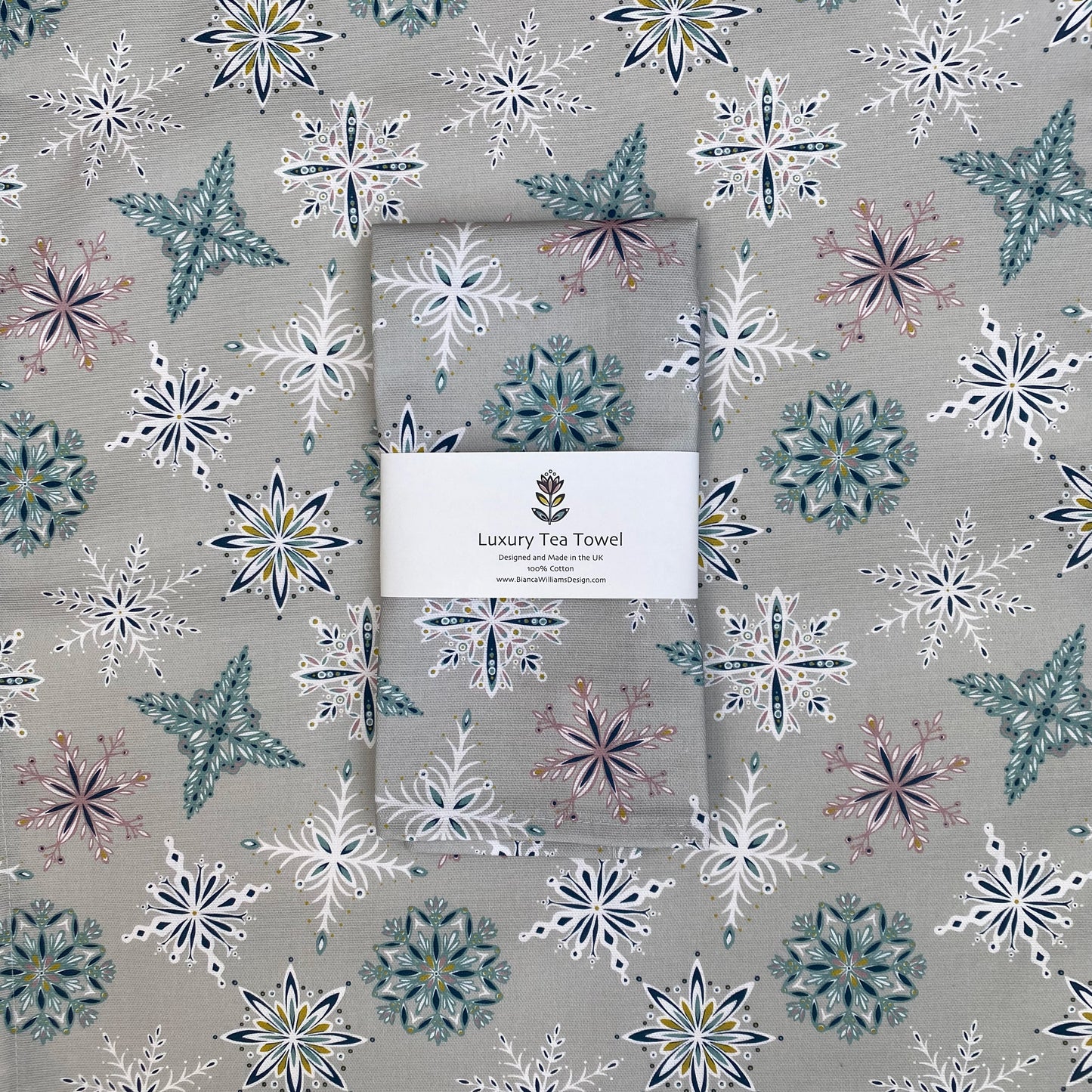 A silver grey snowflake tea towel has been packaged in a branded belly band and placed on an opened out tea towel featuring the same design.  The Snowflake design features white, teal, dusky pink and blue snowflakes on a light grey background.