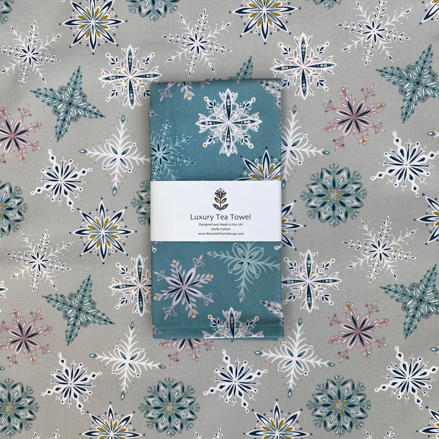 A Blue snowflake tea towel has been packaged in a branded belly band and placed on an opened out tea towel featuring the same design but in a grey colourway.