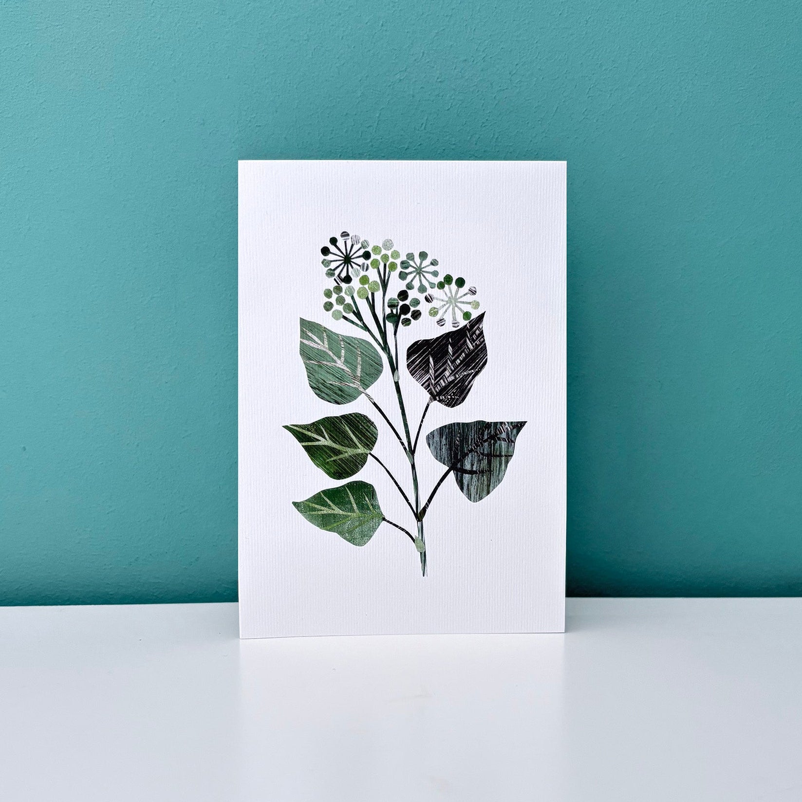 A textured collage of a Green Ivy Leaf on white lightly textured card has been placed on a white shelf against a Teal Wall.