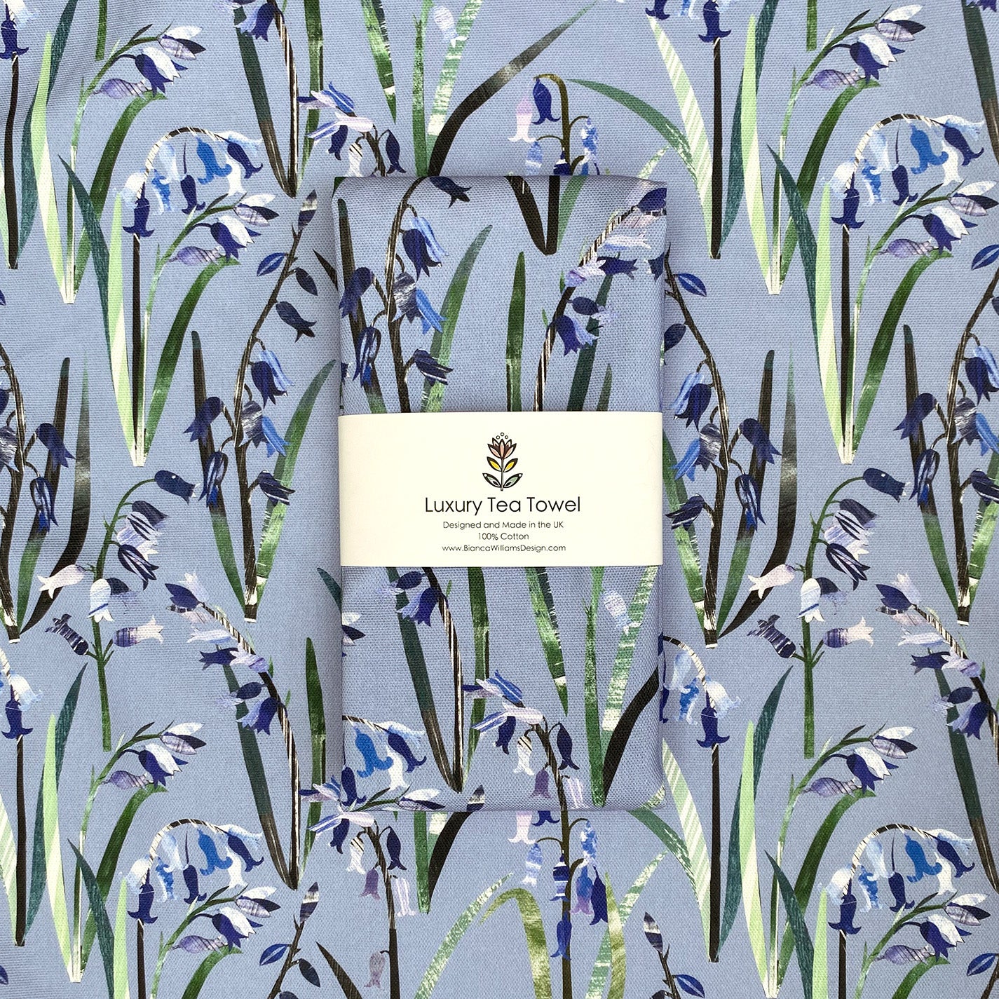 A packaged light blue bluebell tea towel has been folded and packaged in branded belly band and has been placed on an opened out tea towel of the same colour and design.