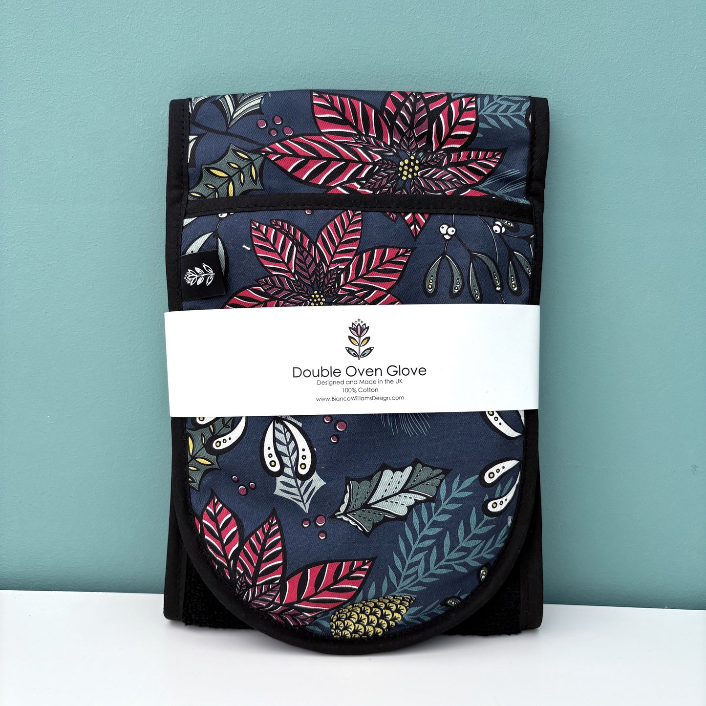 The Navy Poinsettia Oven Gloves have been folded and packaged in a branded belly band and they have been stood on a white shelf against a blue green background. 