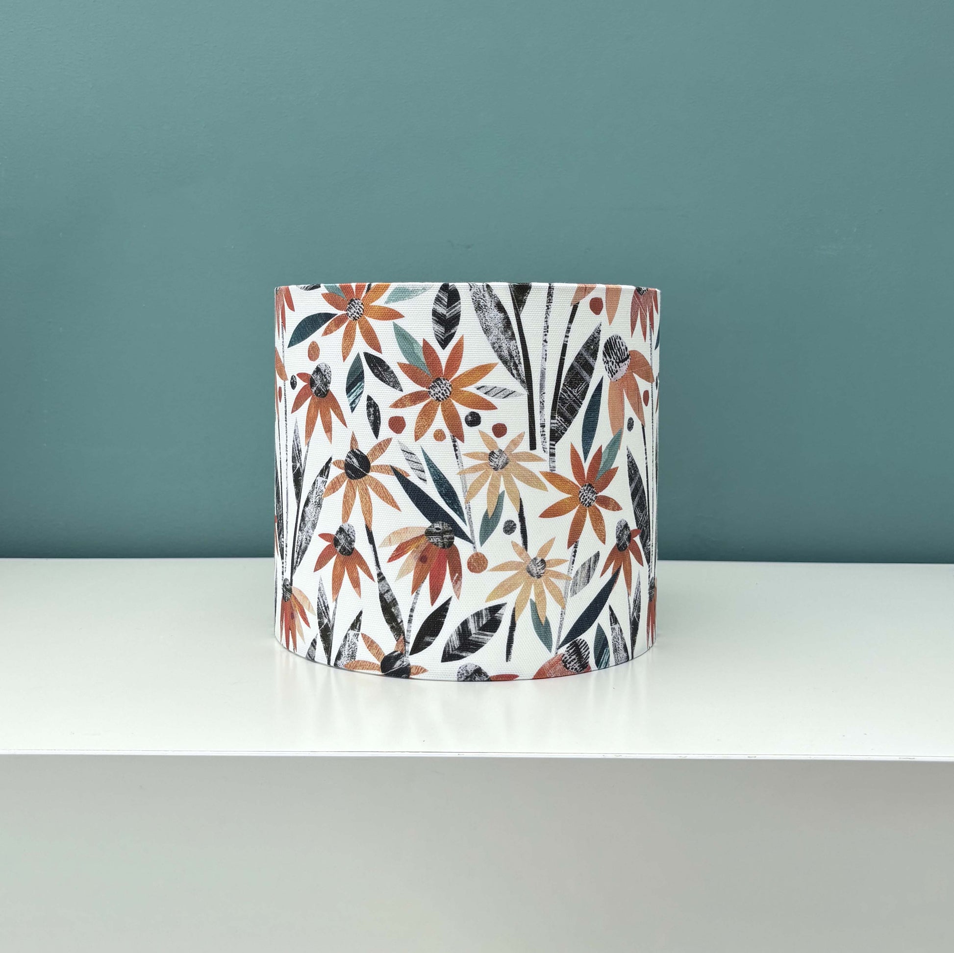 Small Autumn floral lampshade featuring pretty orange rudbeckia flowers with green and dark grey leaves has been placed on a white shelf with a blue green wall behind it.