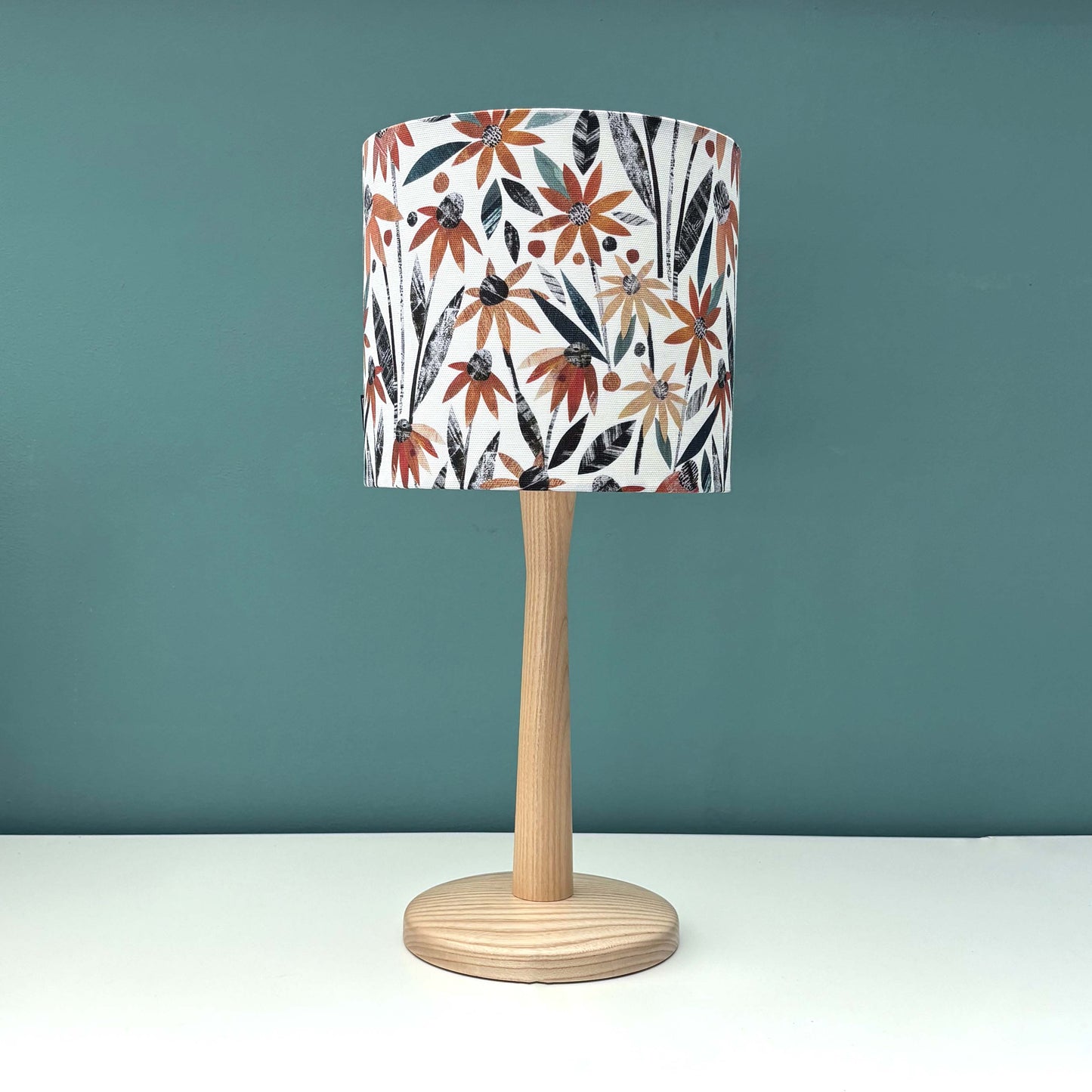 Small Autumn floral lampshade featuring pretty orange rudbeckia flowers with green and dark grey leaves on a white background.  The lampshade has been put on a wooden lamp base and placed on a white shelf with a blue green wall behind it.