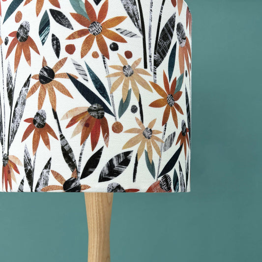 Close up of a Small Autumn floral lampshade featuring pretty orange rudbeckia flowers with green and dark grey leaves on a white background.  The lampshade has been put on a wooden lamp base and has a blue green wall behind it.