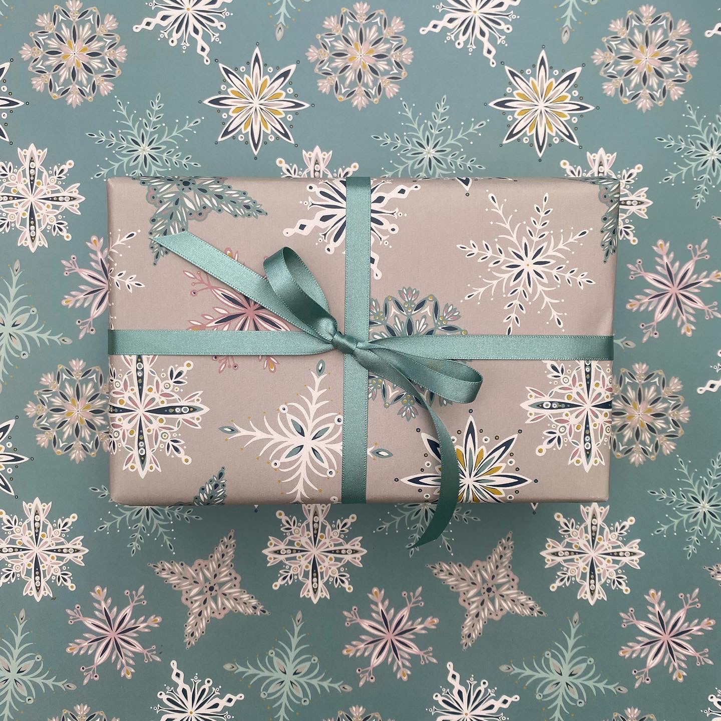 A present has been wrapped in the grey Snowflake gift wrap and has been tied with blue satin ribbon in a bow.  It has been placed on a sheet of the Blue snowflake gift wrap.