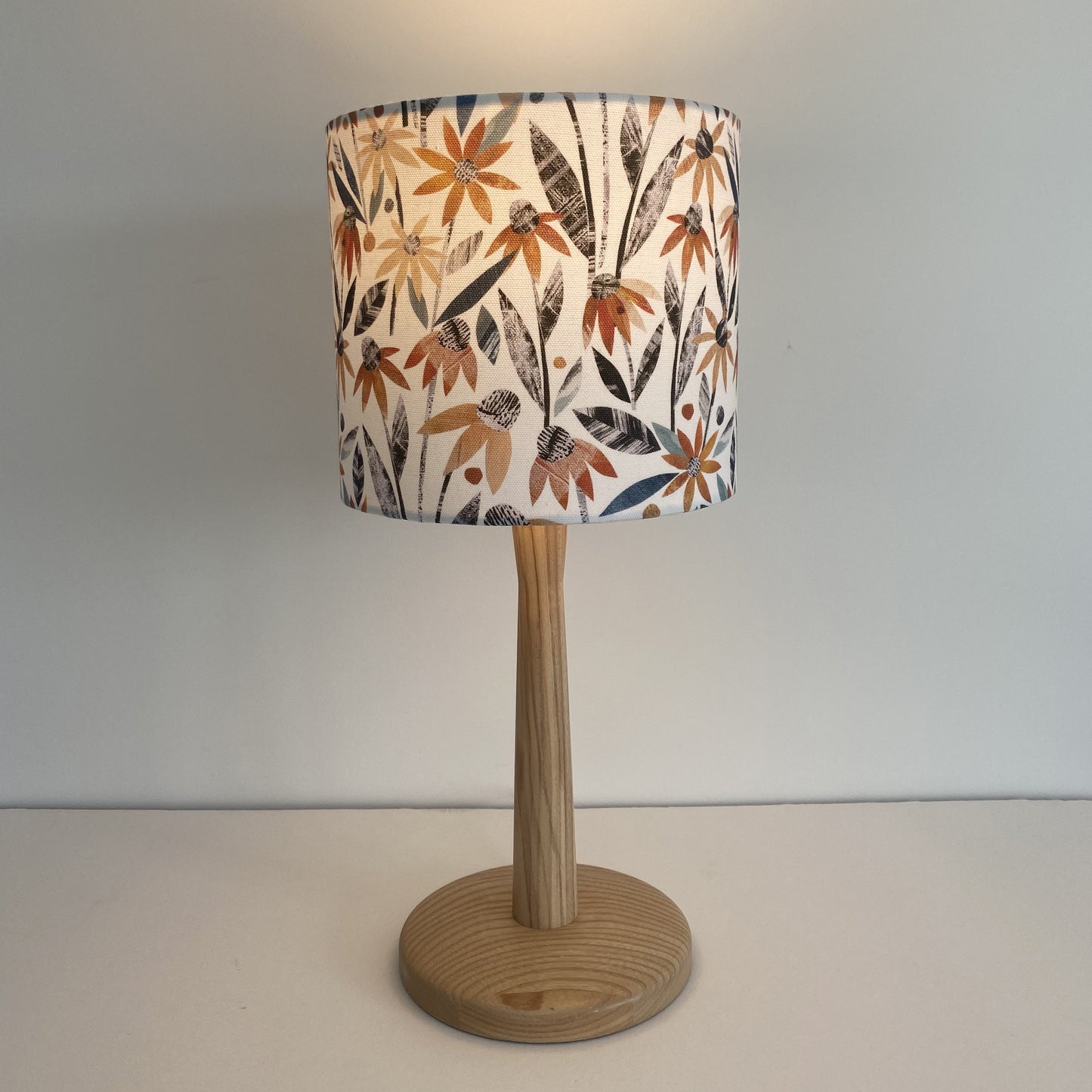 A small Autumn Floral Lampshade is shown here with a wooden lamp base with the light bulb on.