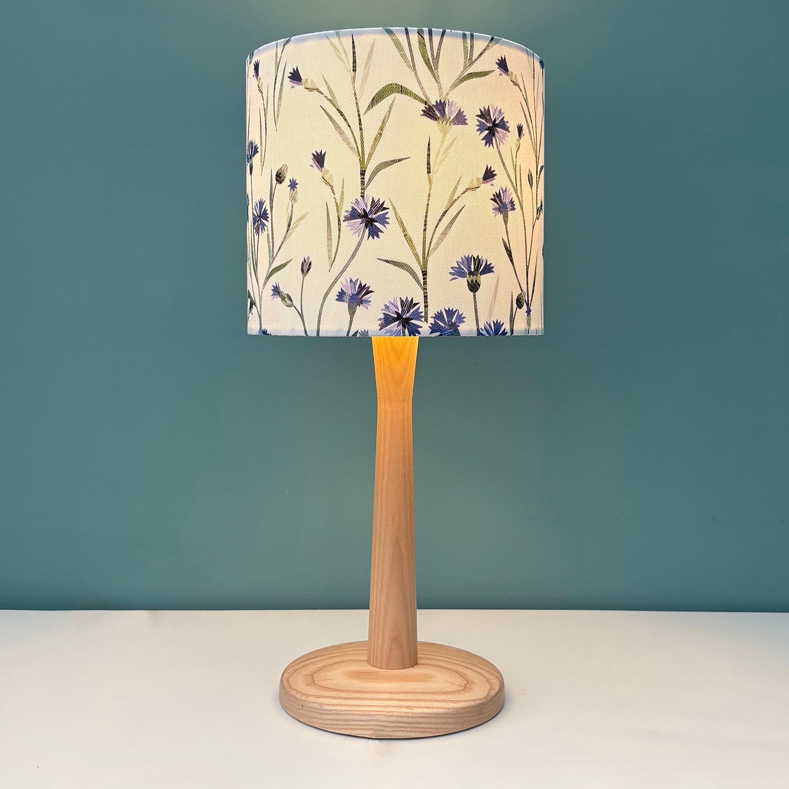 A small floral lampshade featuring blue Cornflowers with green leaves on a white background has been put on a wooden table lamp which has been placed on a white shelf with a blue green wall behind it.