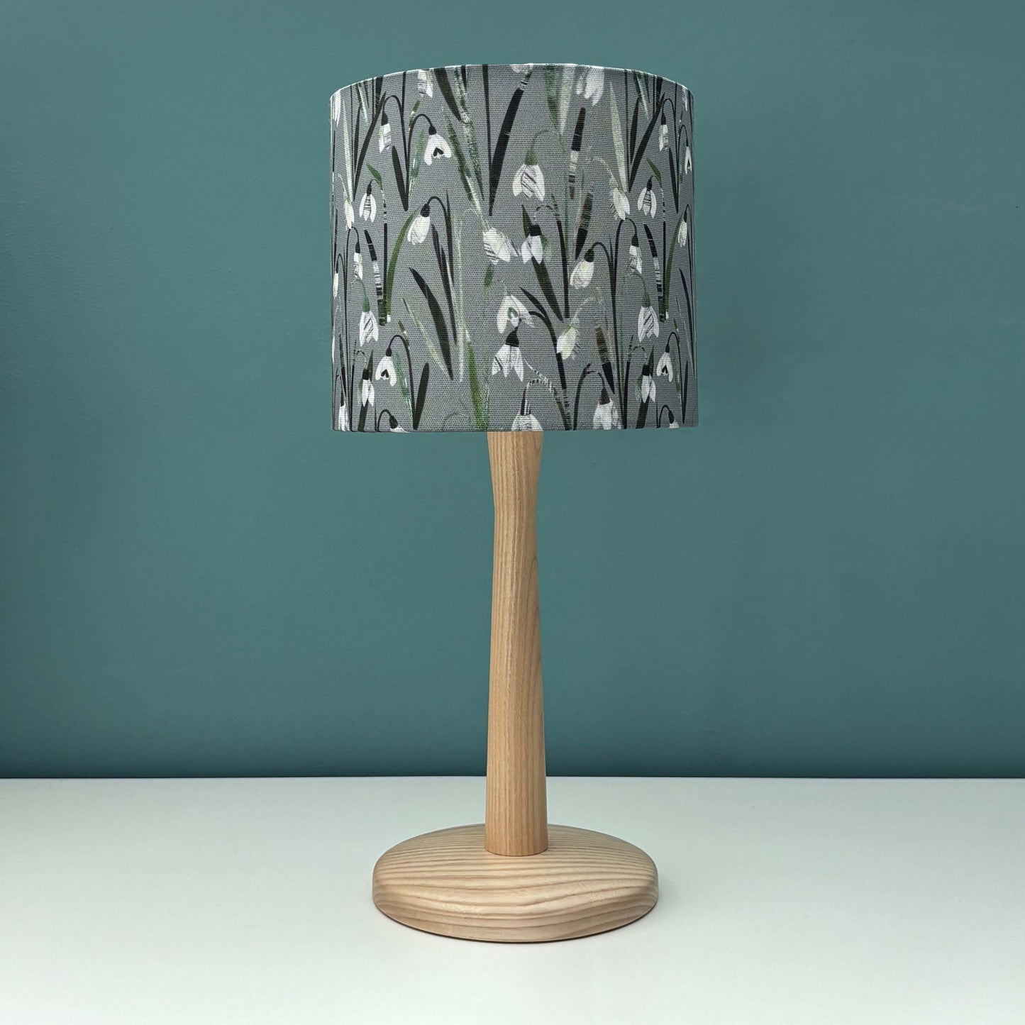 A small Snowdrop lampshade featuring pretty white and cream flowers on a soft sage green background has been put on a wooden lamp base, which has been placed on a white shelf and has a blue green wall behind it.