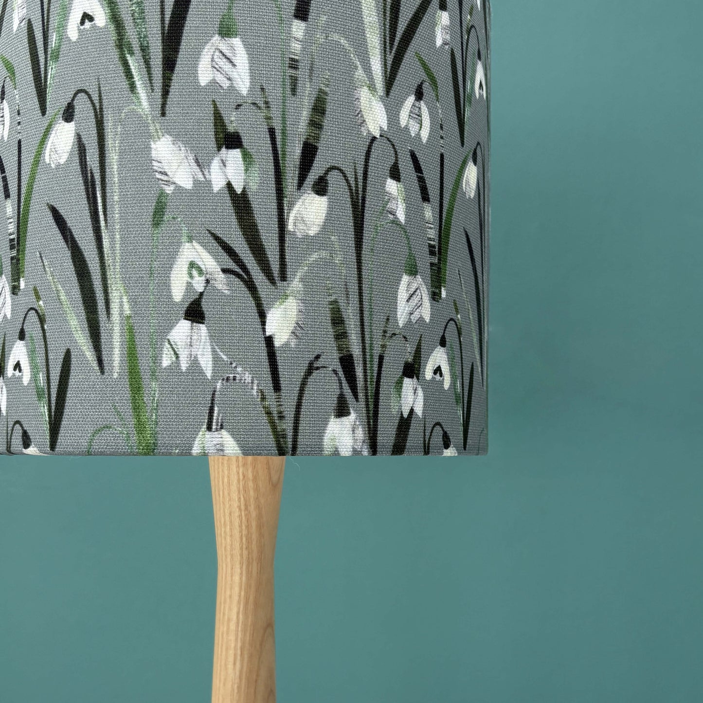 A close up of a small Snowdrop lampshade featuring pretty white and cream flowers on a soft sage green background has been put on a wooden lamp base, which has a blue green wall behind it.