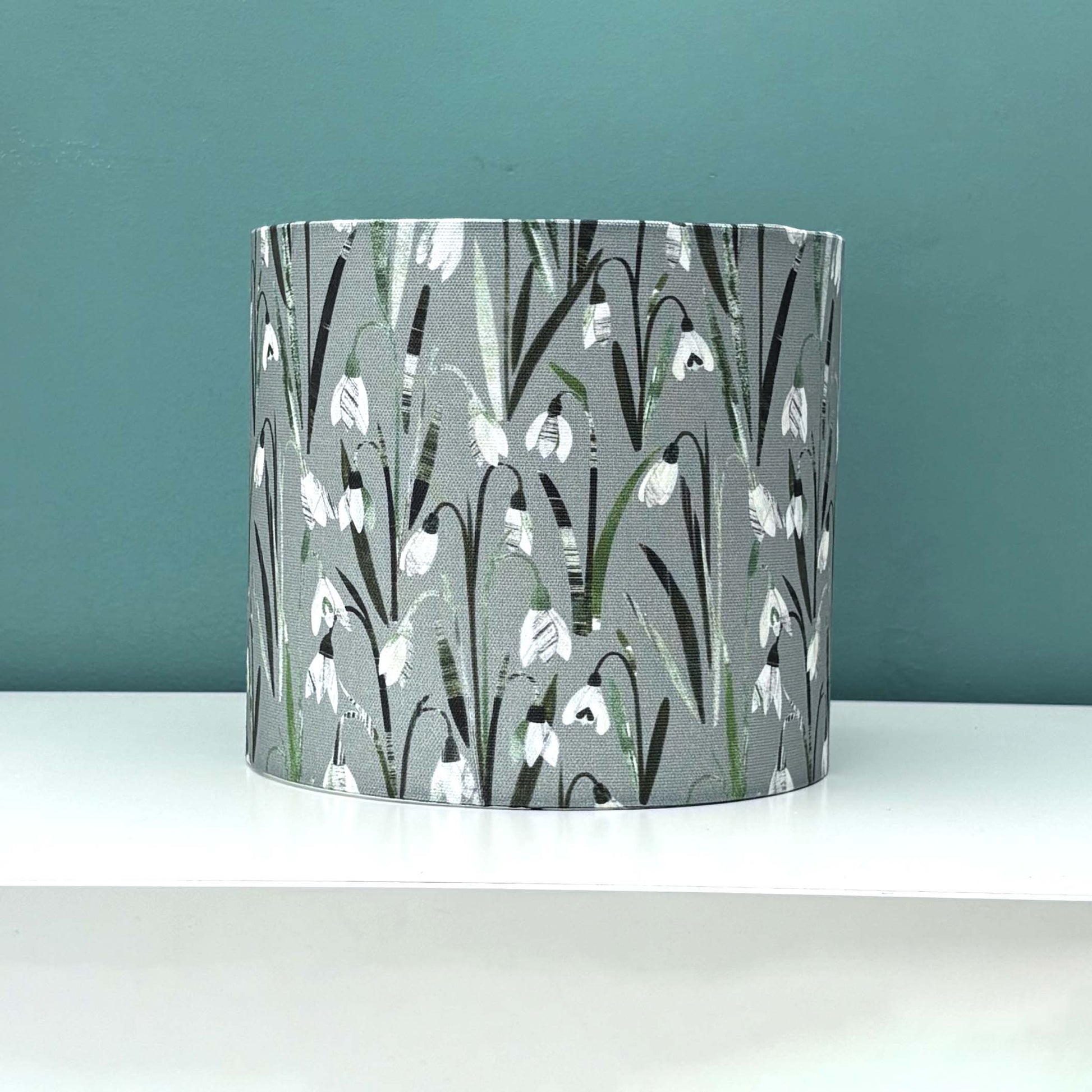 A small Snowdrop lampshade featuring pretty white and cream flowers on a soft sage green background has been placed on a white shelf and has a blue green wall behind it.