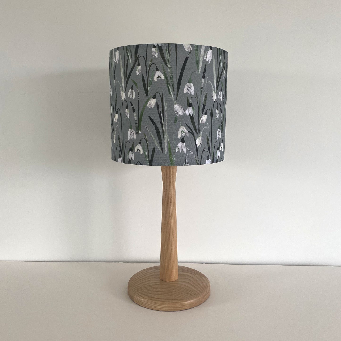 Small Lampshade featuring pretty snowdrop flowers with green leaves on a green/grey textured background.  The snowdrops lampshade has a light wooden lamp base.