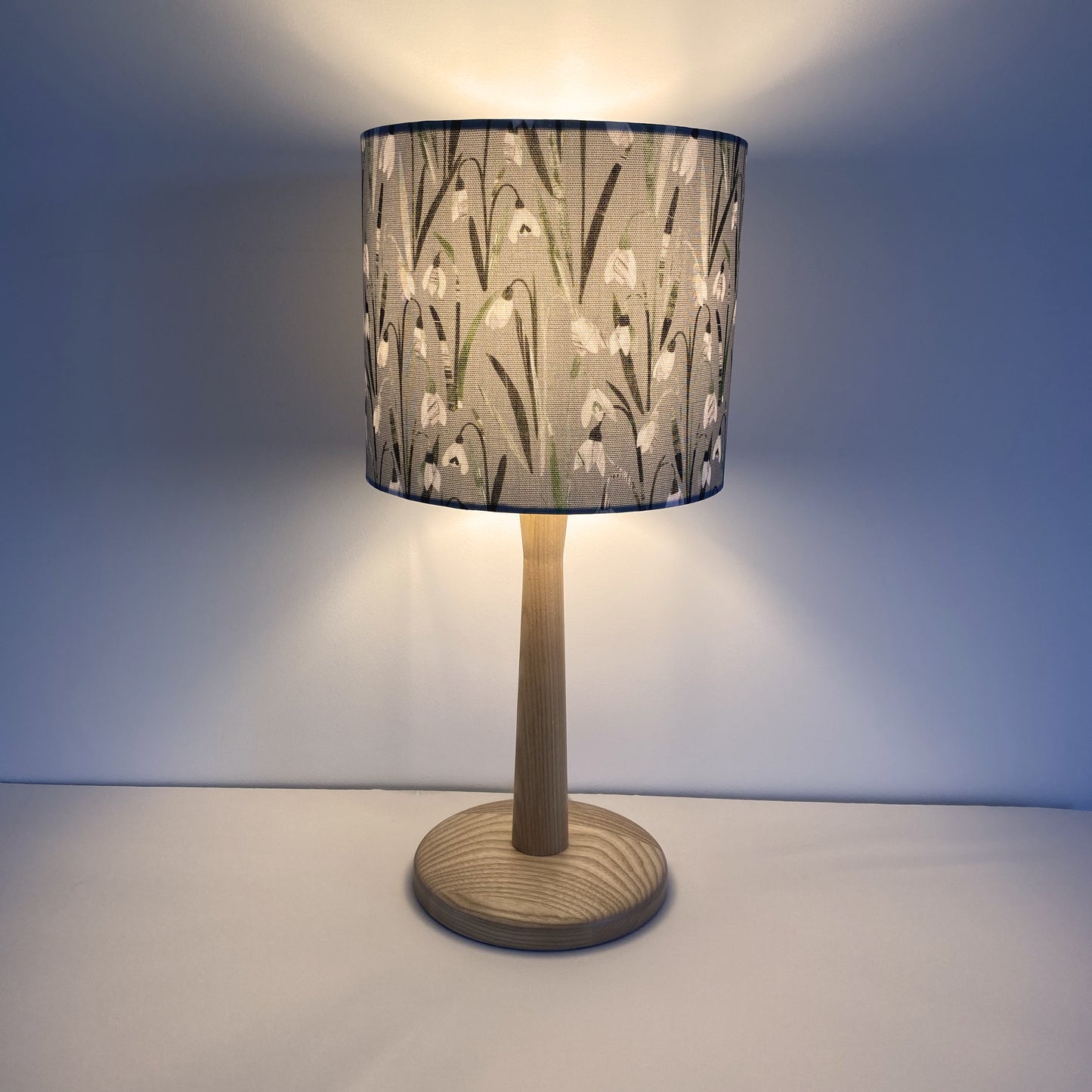 Small Lampshade featuring pretty snowdrop flowers with green leaves on a green/grey textured background.  The snowdrops lampshade has a light wooden lamp base and is shown with the light on.