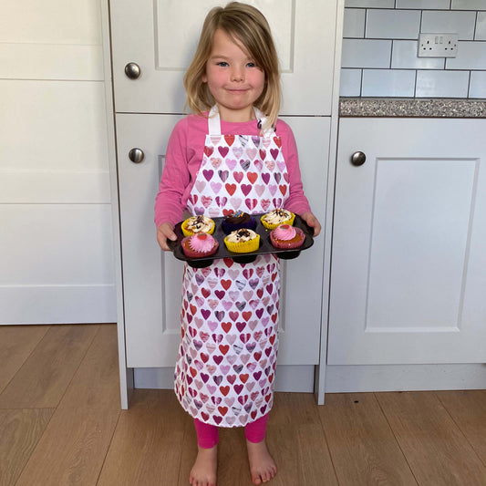 A young girl stands barefoot in a kitchen holding a tray of cupcakes, wearing a pink long-sleeve shirt and a white apron with a heart pattern.