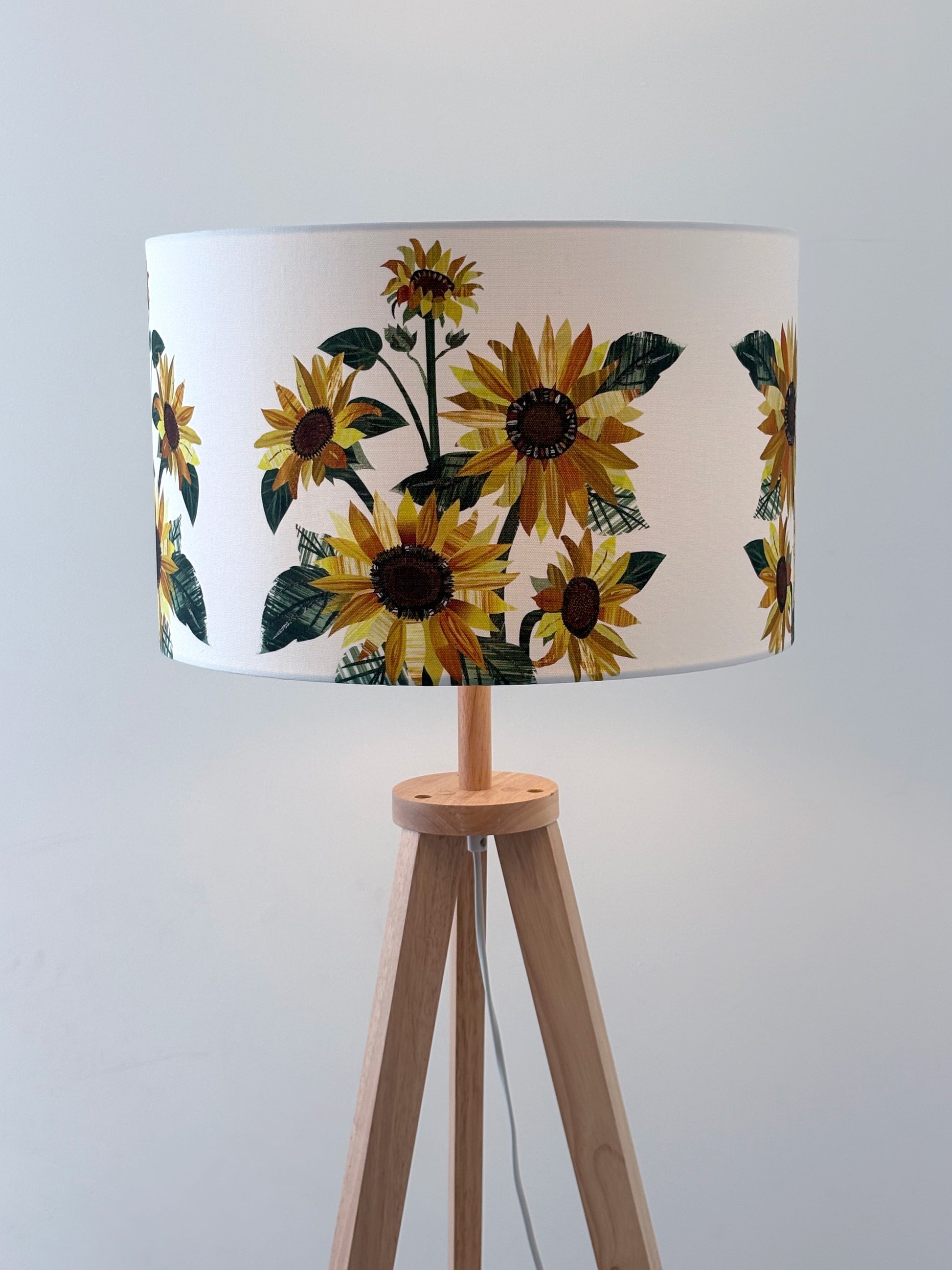 A large Sunflower lampshade has been placed on a wooden lamp base and it is against a pale grey background.