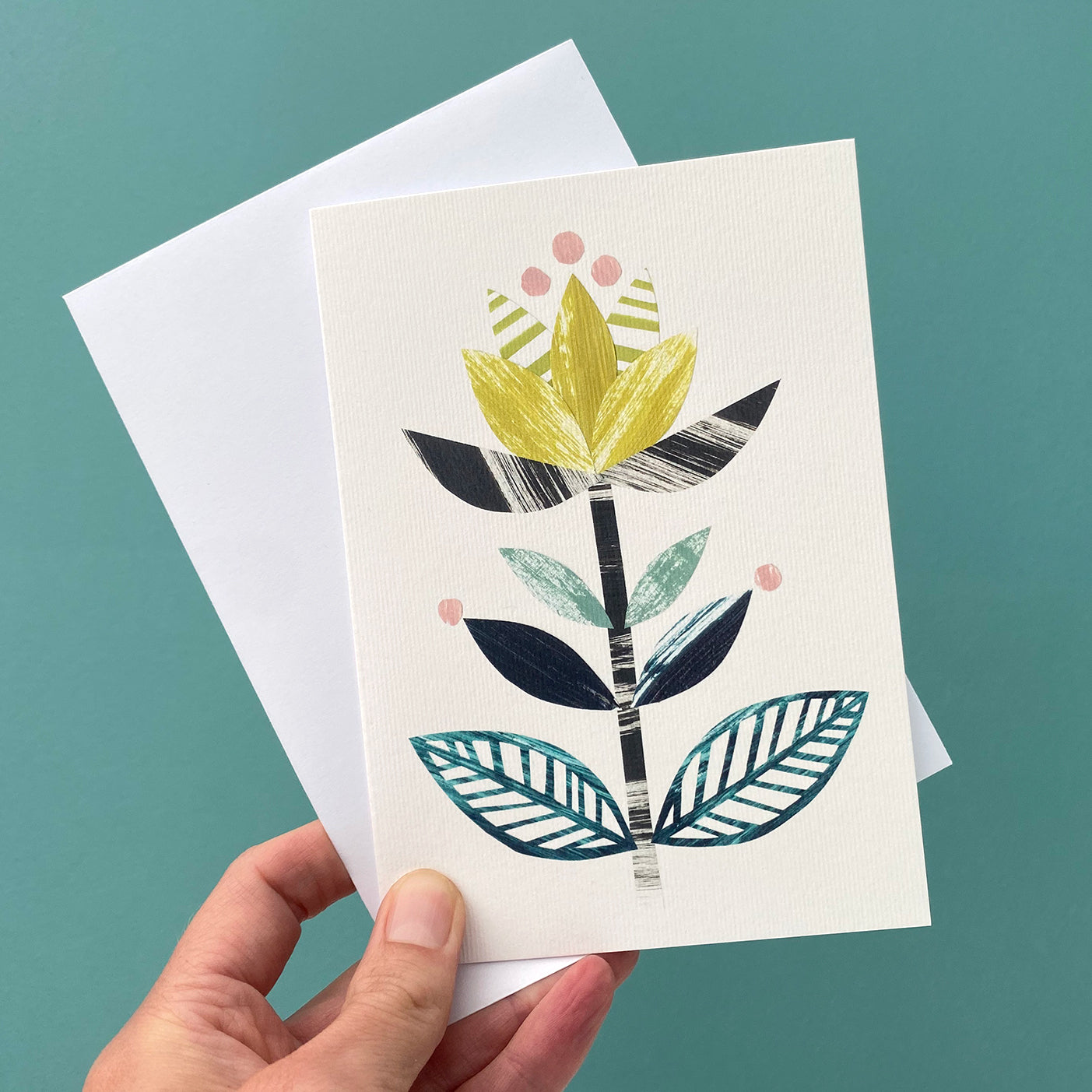 A card featuring a stylised flower motif in yellow, greens and blacks on a white background is being held with a white envelope against a blue green background.