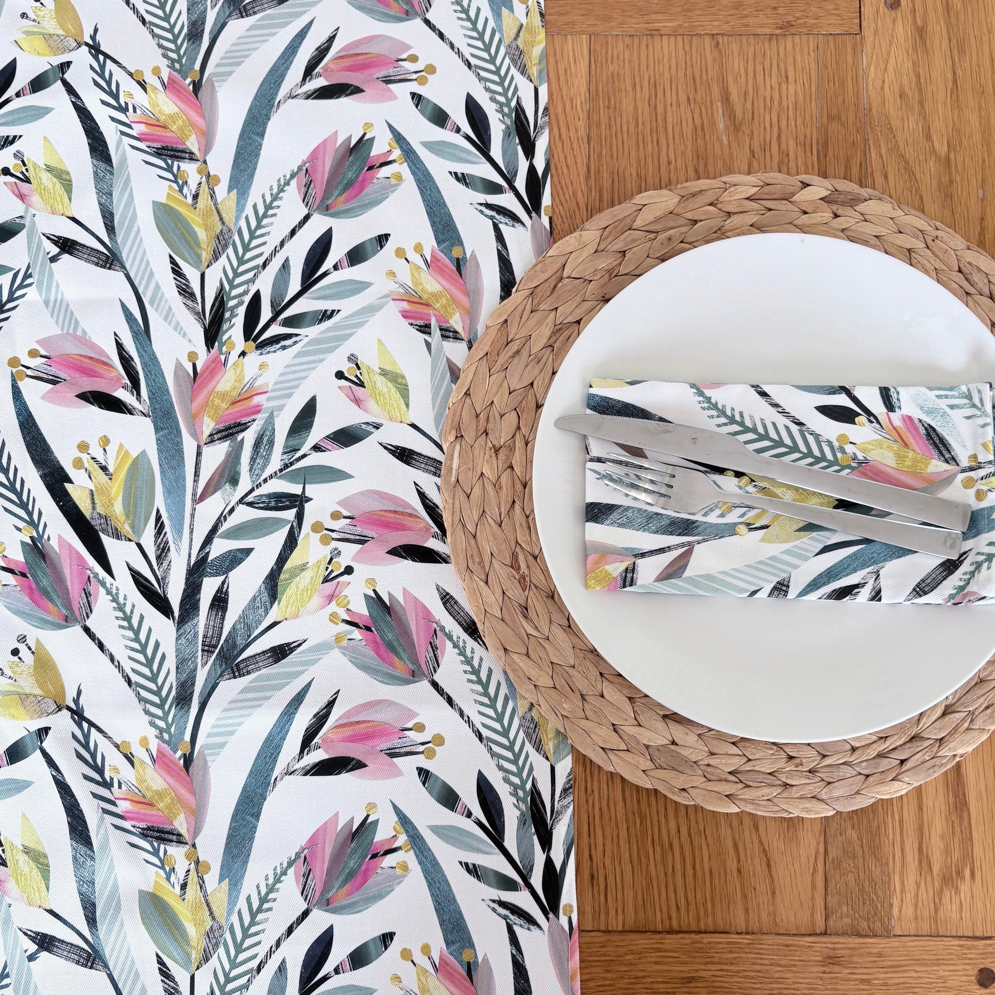 A colourful tulip Floral patterned table runner on a wooden table with woven placemats, with a white plate and matching floral cotton napkin on it.