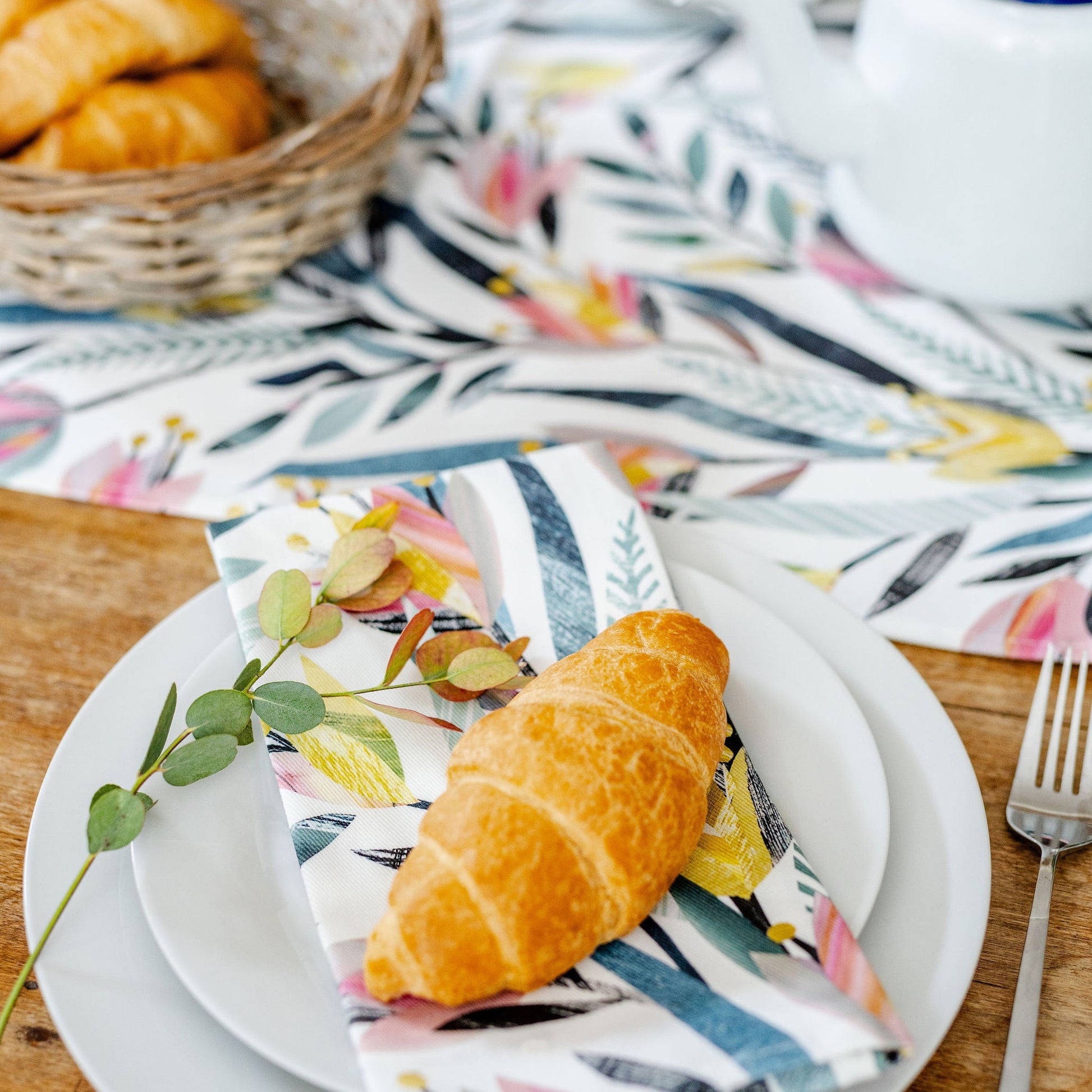 Breakfast setting with a croissant on a colourful tulips napkin, teapot, and basket of bread.