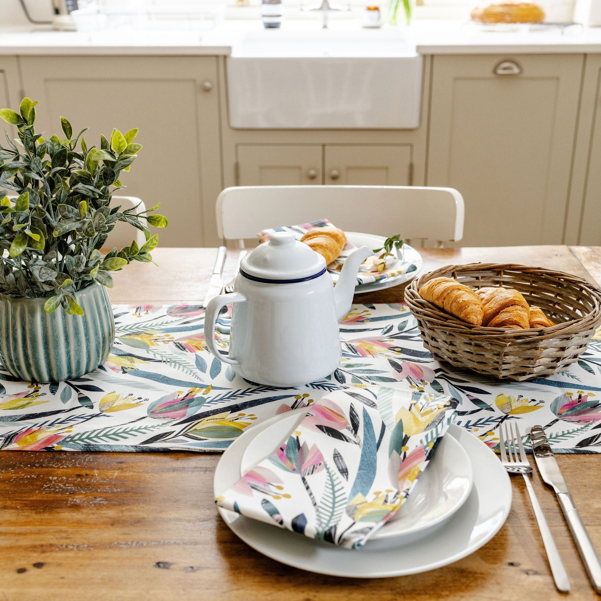 Dining table set with colourful tulip floral table runner and matching napkins with a  teapot, plates, and basket of bread on it in a cream kitchen.