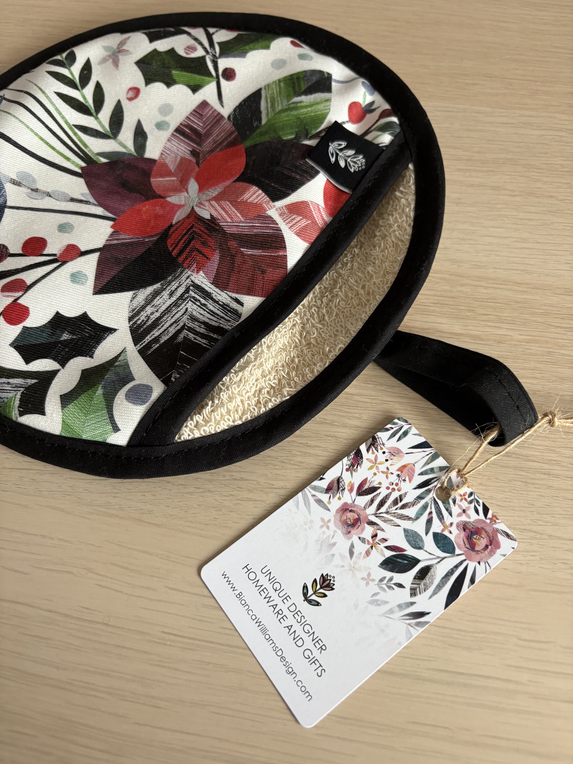 A Winter Floral Pot holder has been placed on a wooden table, you can see see the brand label sewn into the pocket for the hand and a swing tag has been attached with twine.