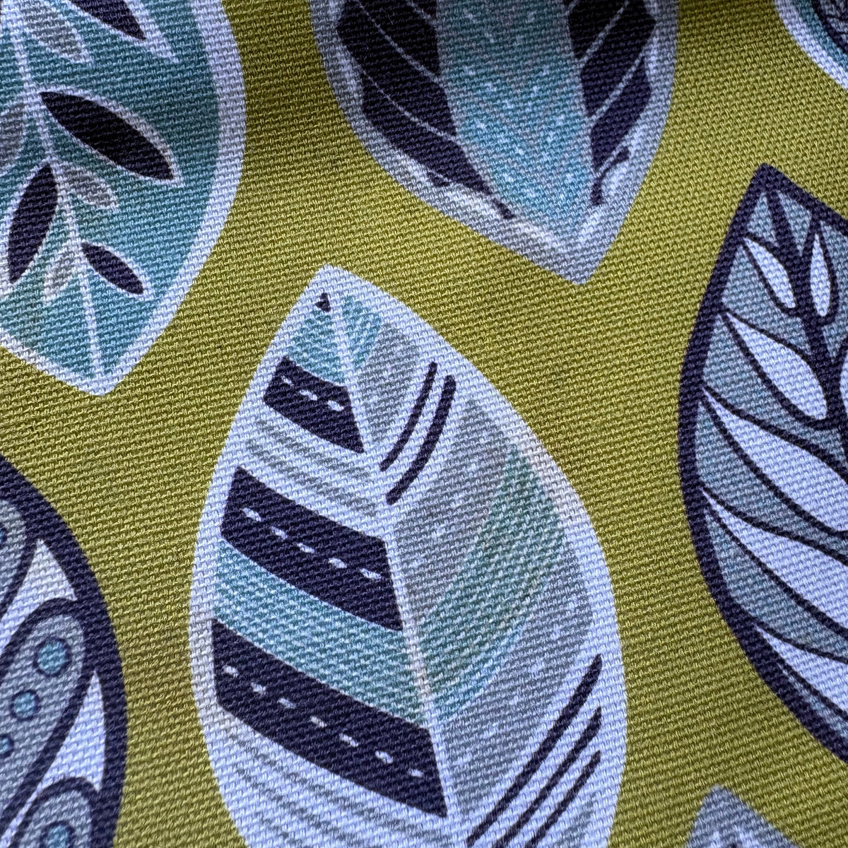 A close-up of a woven fabric with a repeating leaf pattern in teal, navy, and white on a mustard yellow background.
