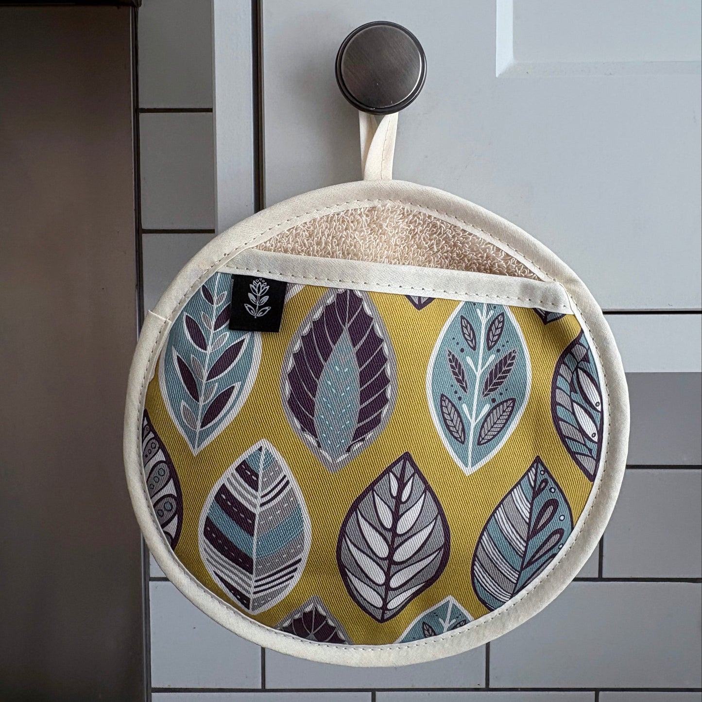 A round potholder with a mustard yellow background and a pattern of stylised leaves in blue, purple, and grey, hanging from a black hook on a pale grey panelled kitchen door.